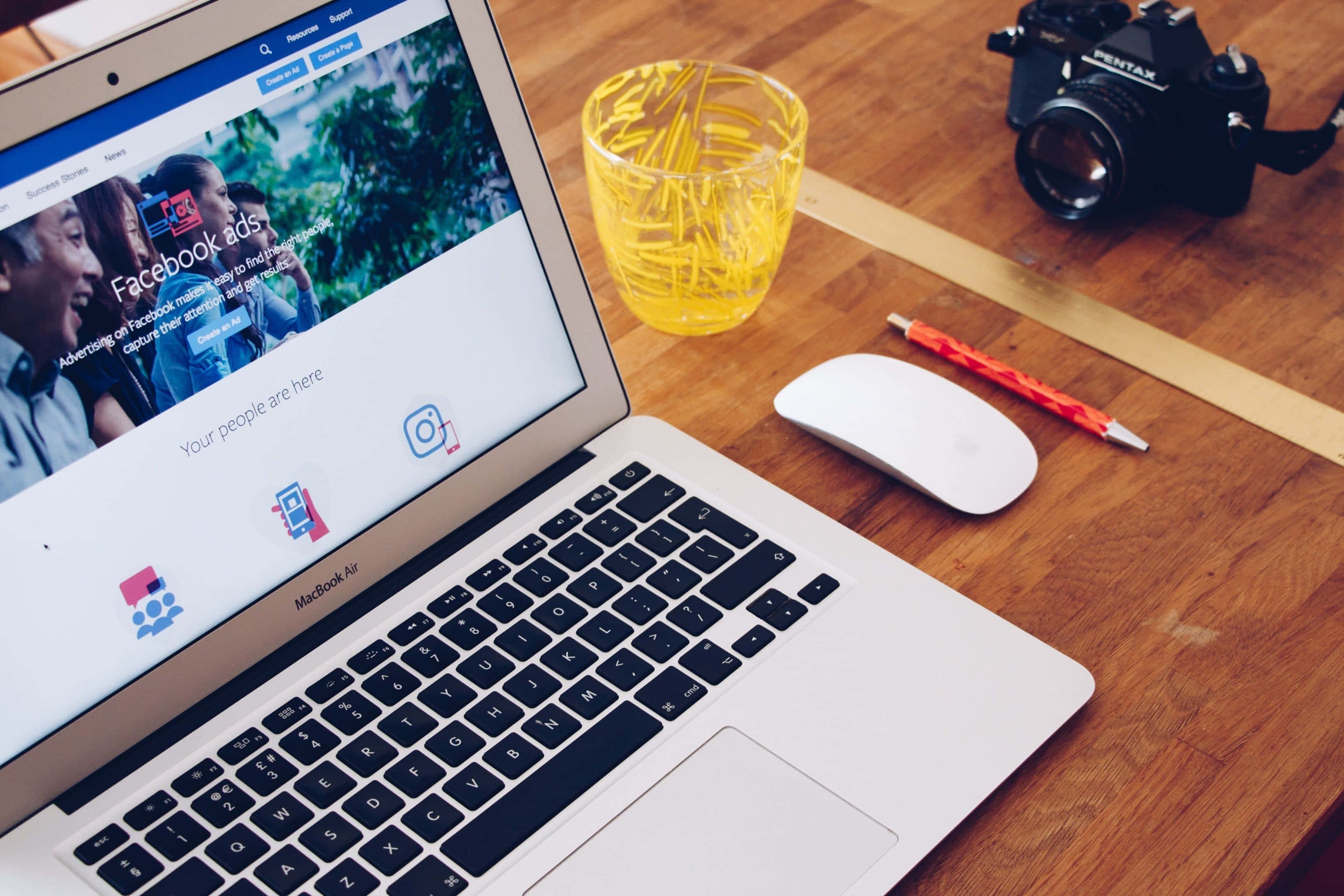 Telemarketing 桌布 壁紙 — 一部銀色 MacBook Air 在木桌上顯示 Facebook 廣告，旁邊有黃色玻璃杯、紅色筆、白色滑鼠和黑色 Pentax 相機。