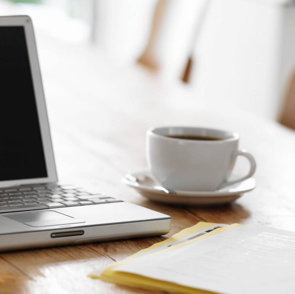 Best Laptop 桌布 壁紙 — 一部銀色手提電腦和一個白色咖啡杯放在木桌上，旁邊有黃色信封和紙張，在柔和自然光下。