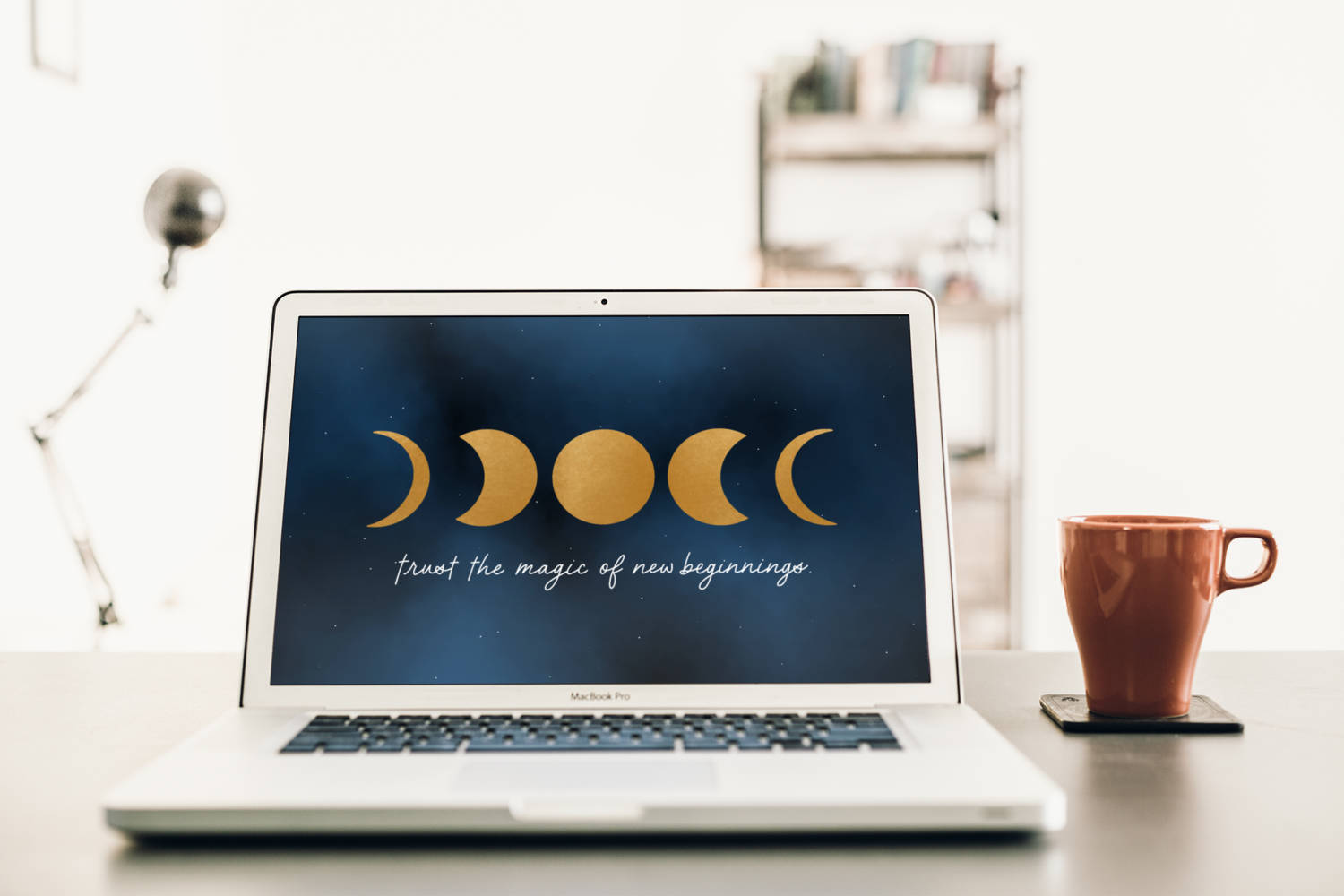Working From Home 桌布 壁紙 — 一部銀色手提電腦在簡約的桌子上，旁邊有一個陶土杯，顯示金色的月相和鼓舞人心的文字。
