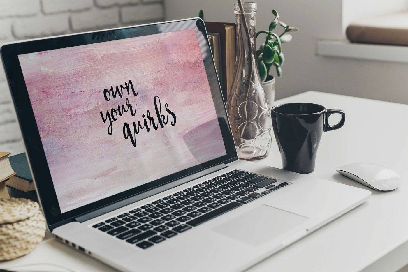 Laptop 桌布 壁紙 — 一部銀色手提電腦在粉紅色水彩背景上顯示「own your quirks」，旁邊有一個黑色馬克杯和一個裝有綠色植物的玻璃花瓶，放在白色桌子上。