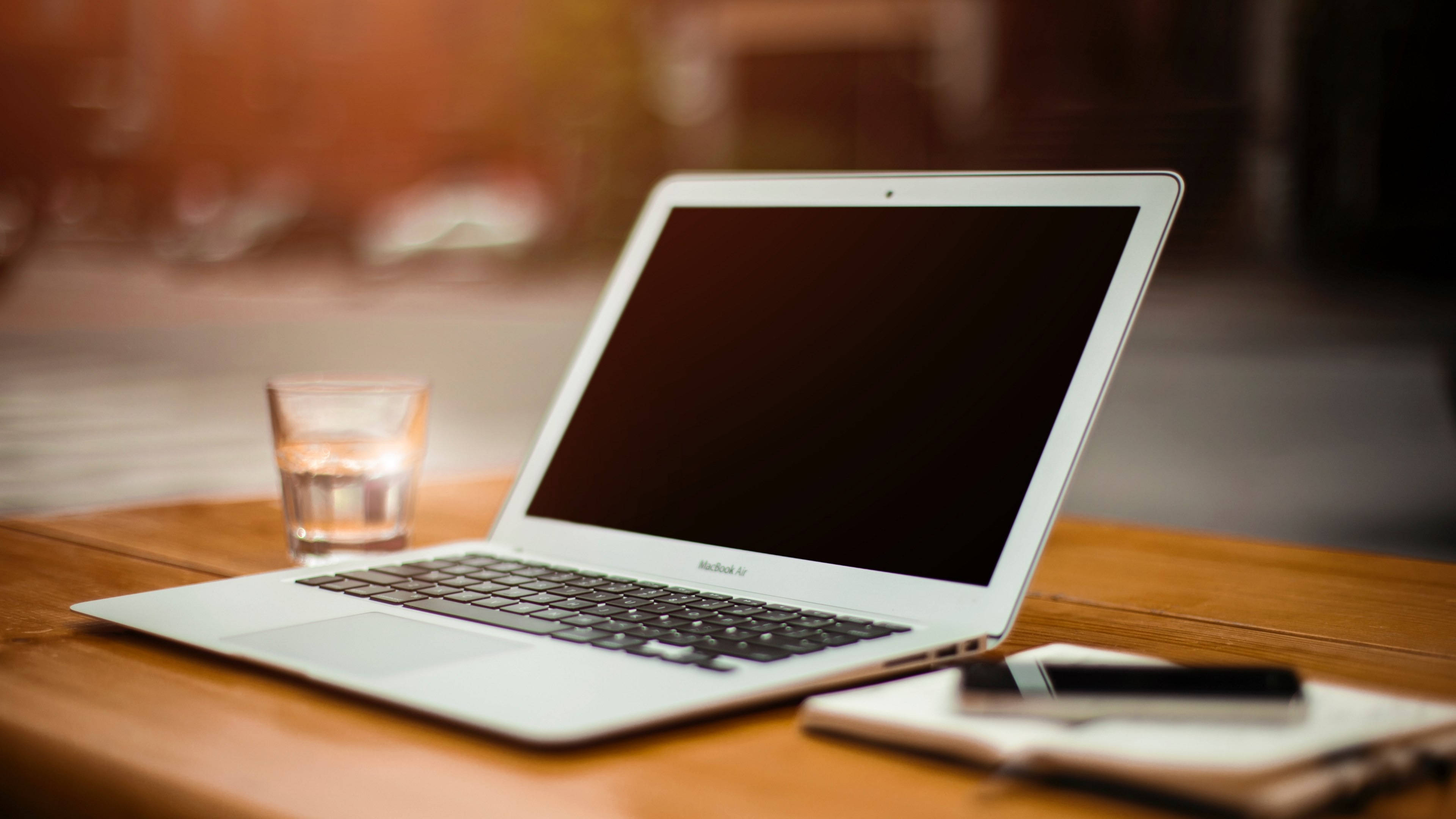macbook 桌布 壁紙 — 一部銀色手提電腦打開放在木桌上，旁邊有一杯水和一本帶筆的筆記本。
