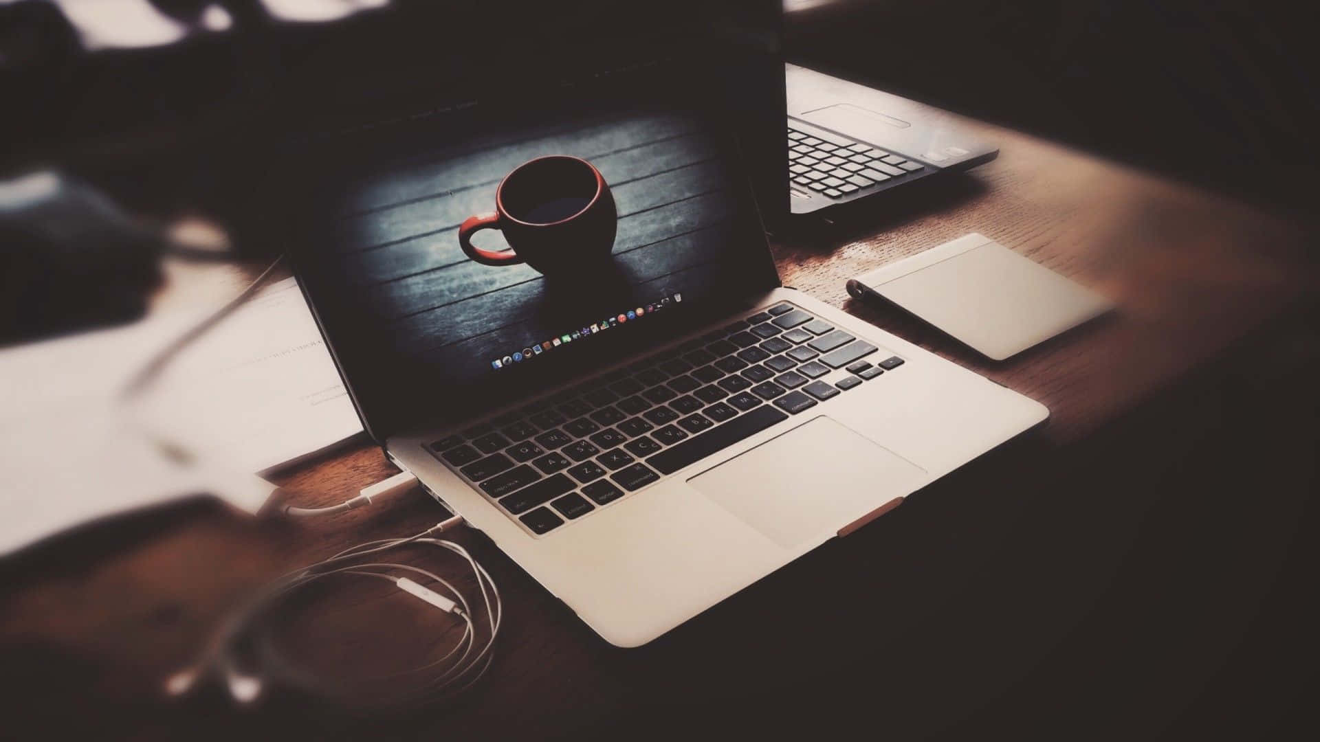 Best Laptop 桌布 壁紙 — 一部銀色手提電腦放在木桌上，顯示咖啡杯壁紙，周圍有電線和筆記本，在溫暖昏暗的燈光下。