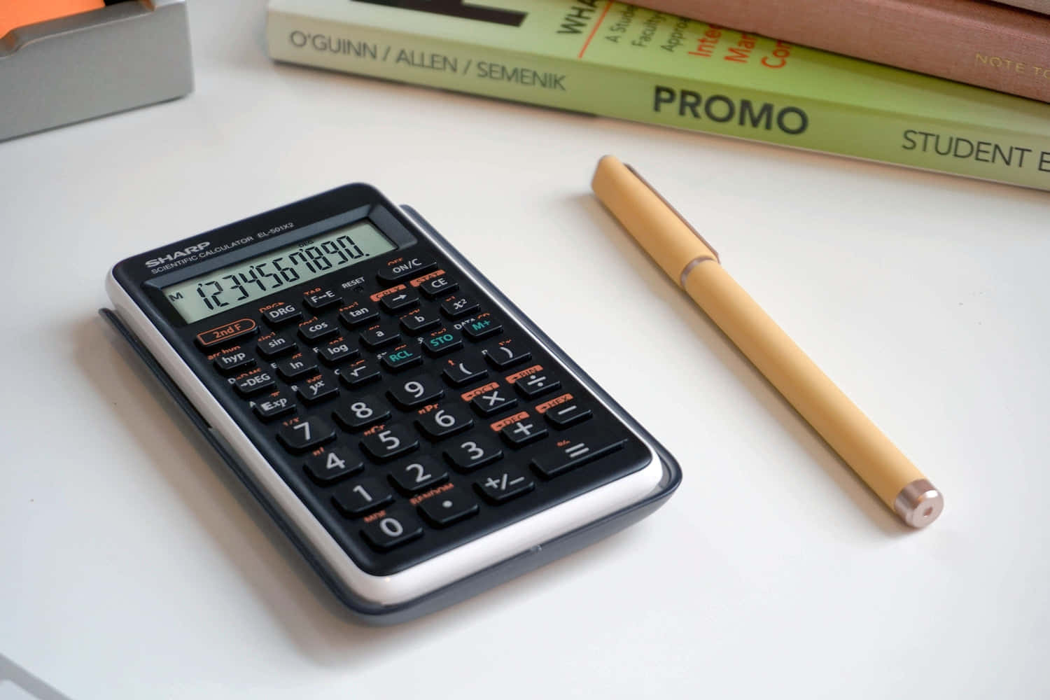 Calculator 桌布 壁紙 — 一部顯示1234567890的黑色科學計算機旁邊放著一支米色原子筆，白色書桌上有教科書，背景柔和。