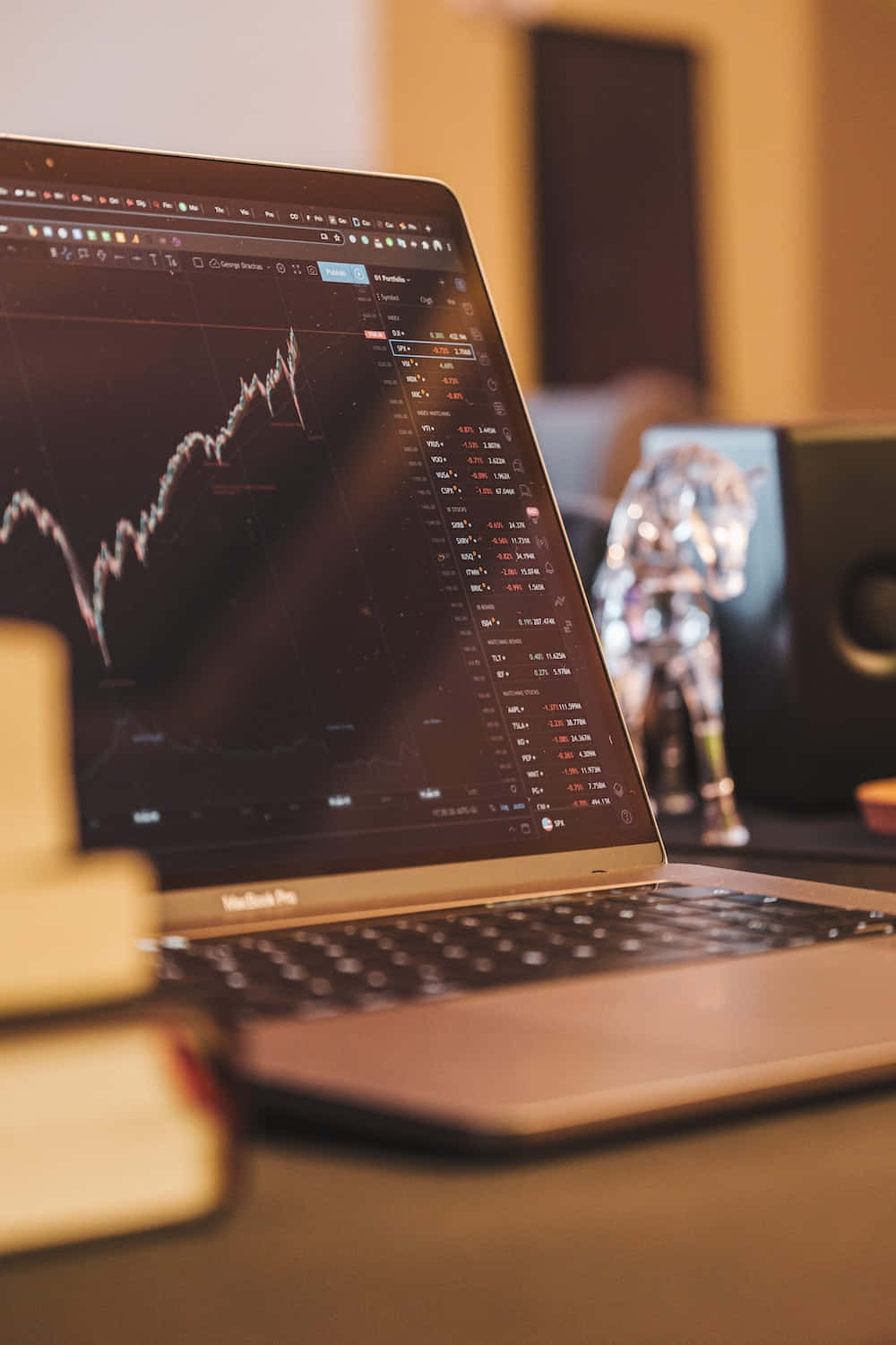 Stock Market 桌布 壁紙 — 一部顯示波動股票圖表的筆記型電腦放在木桌上，旁邊有書本和一個金屬公仔，在溫暖的室內燈光下。
