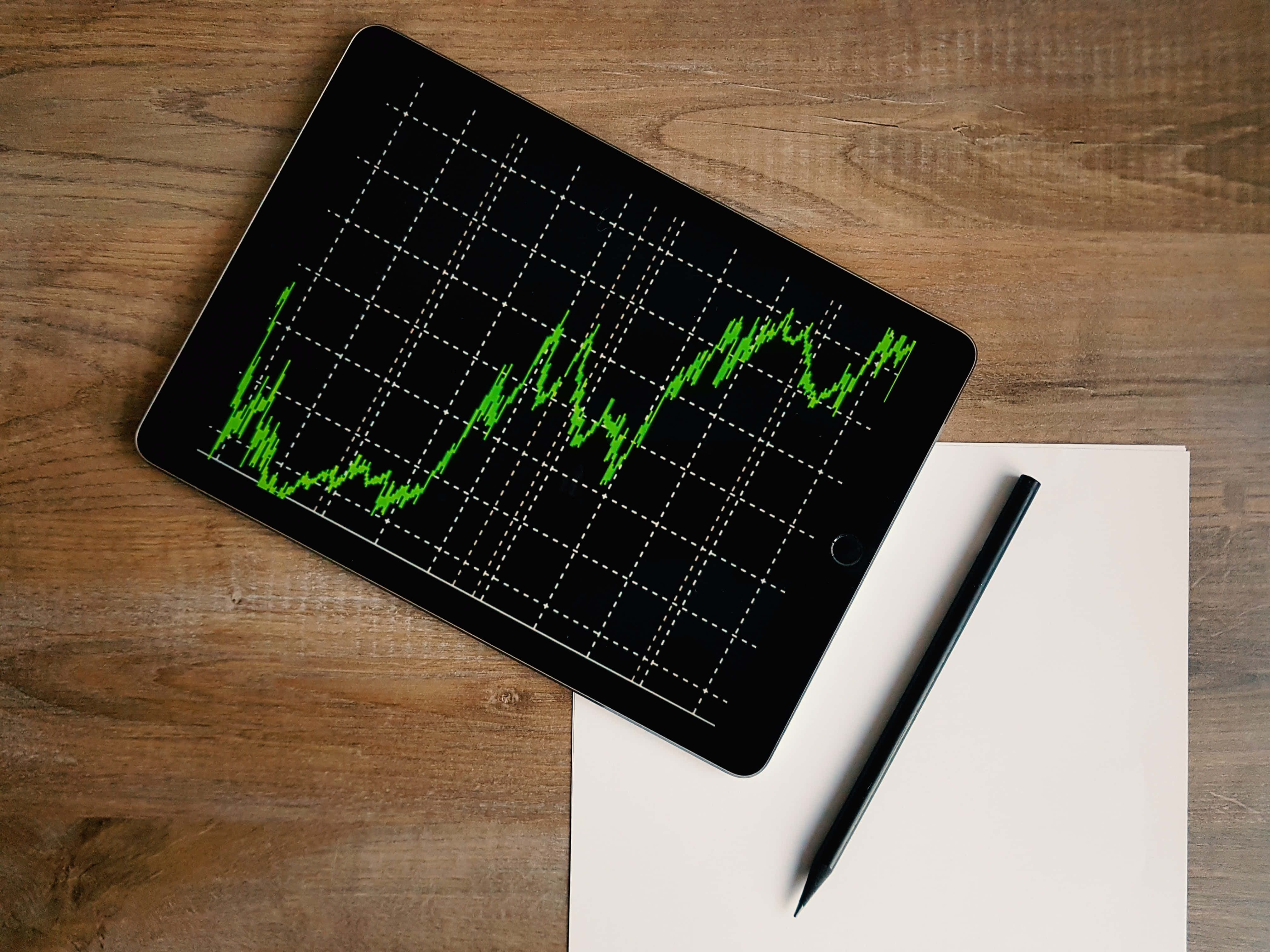 Stock Market 桌布 壁紙 — 一部顯示鮮綠色股票圖表的平板電腦放在木桌上，旁邊有一個空白筆記本和黑色筆。