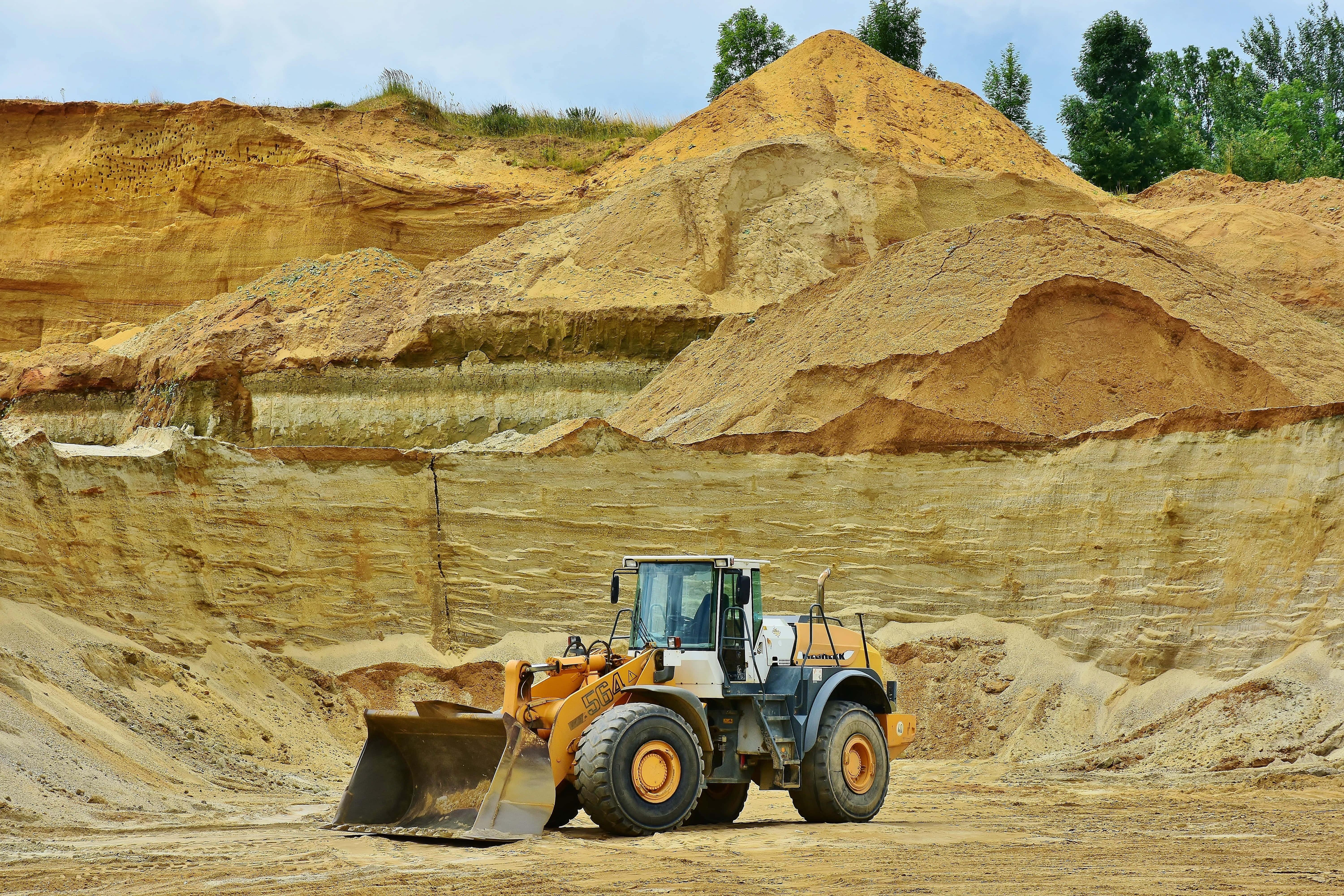 Mining 桌布 壁紙 — 一部黃色嘅前裝載機喺廣闊嘅露天採石場，喺晴朗嘅藍天下，準備好喺層狀泥土同稀疏嘅綠樹之間工作。