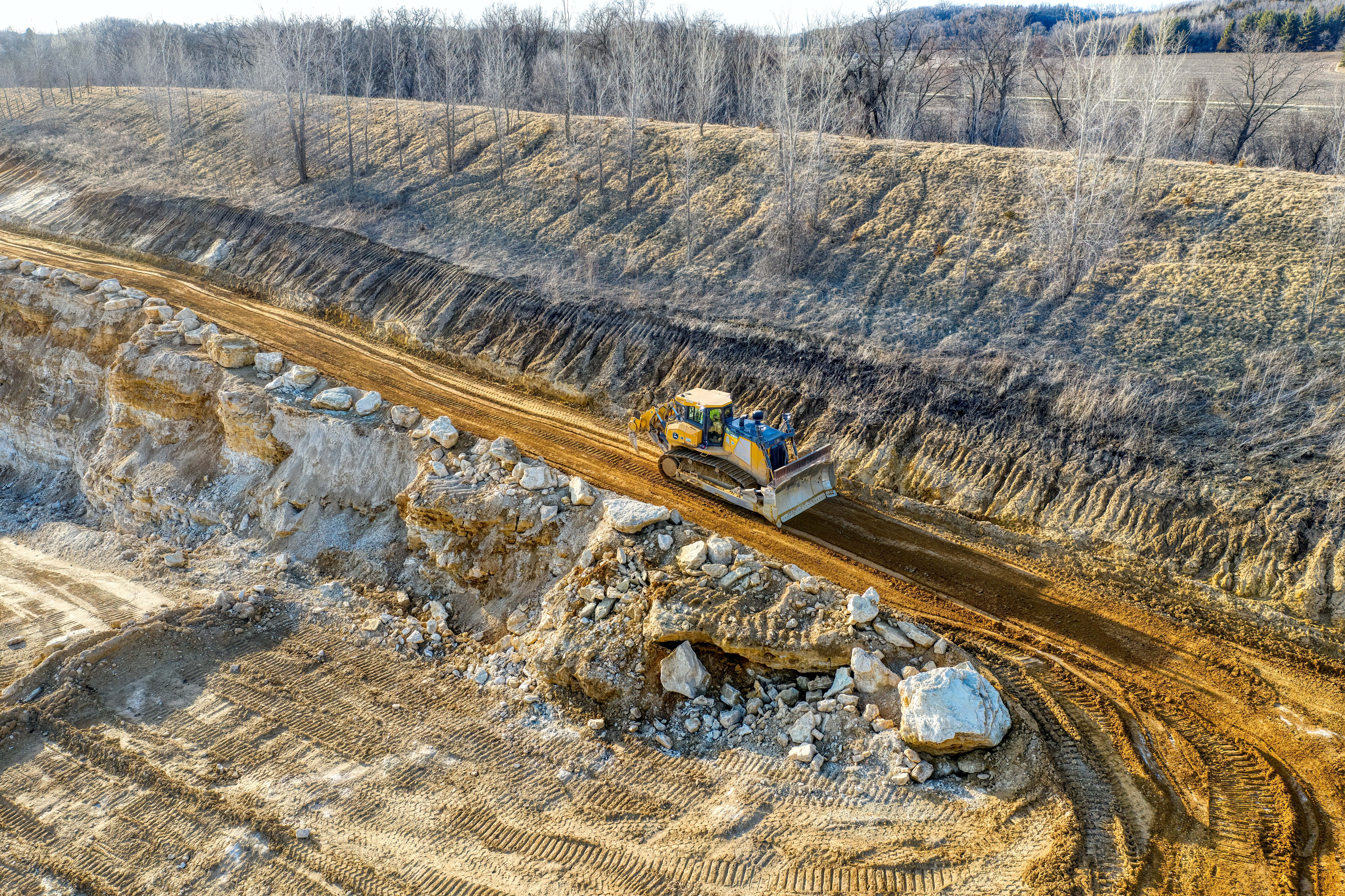 Mining 桌布 壁紙 — 一部黃色推土機在光禿禿的採石場中，沿著崎嶇的泥土路推動泥土，周圍是沒有葉子的樹木，陽光普照。