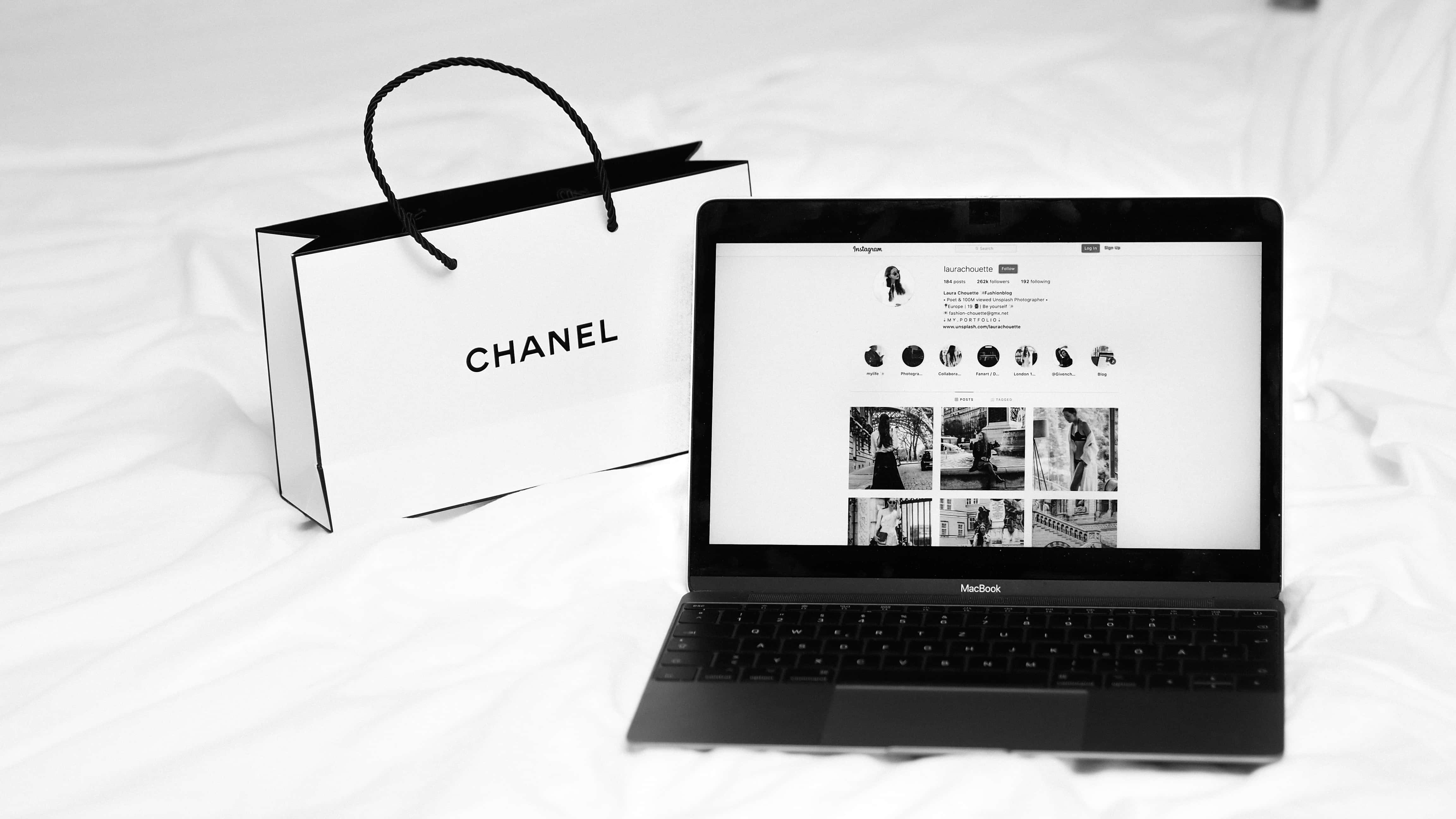 黑白美學 桌布 壁紙 — 一部黑白 MacBook 在皺巴巴的白色床單上顯示 Instagram 個人資料，旁邊放著一個 Chanel 購物袋。