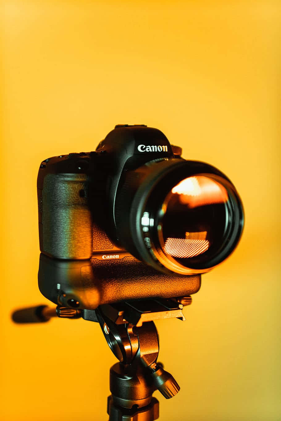 Dslr Camera 桌布 壁紙 — 一部黑色Canon DSLR相機安裝在三腳架上，背景為溫暖的金黃色。