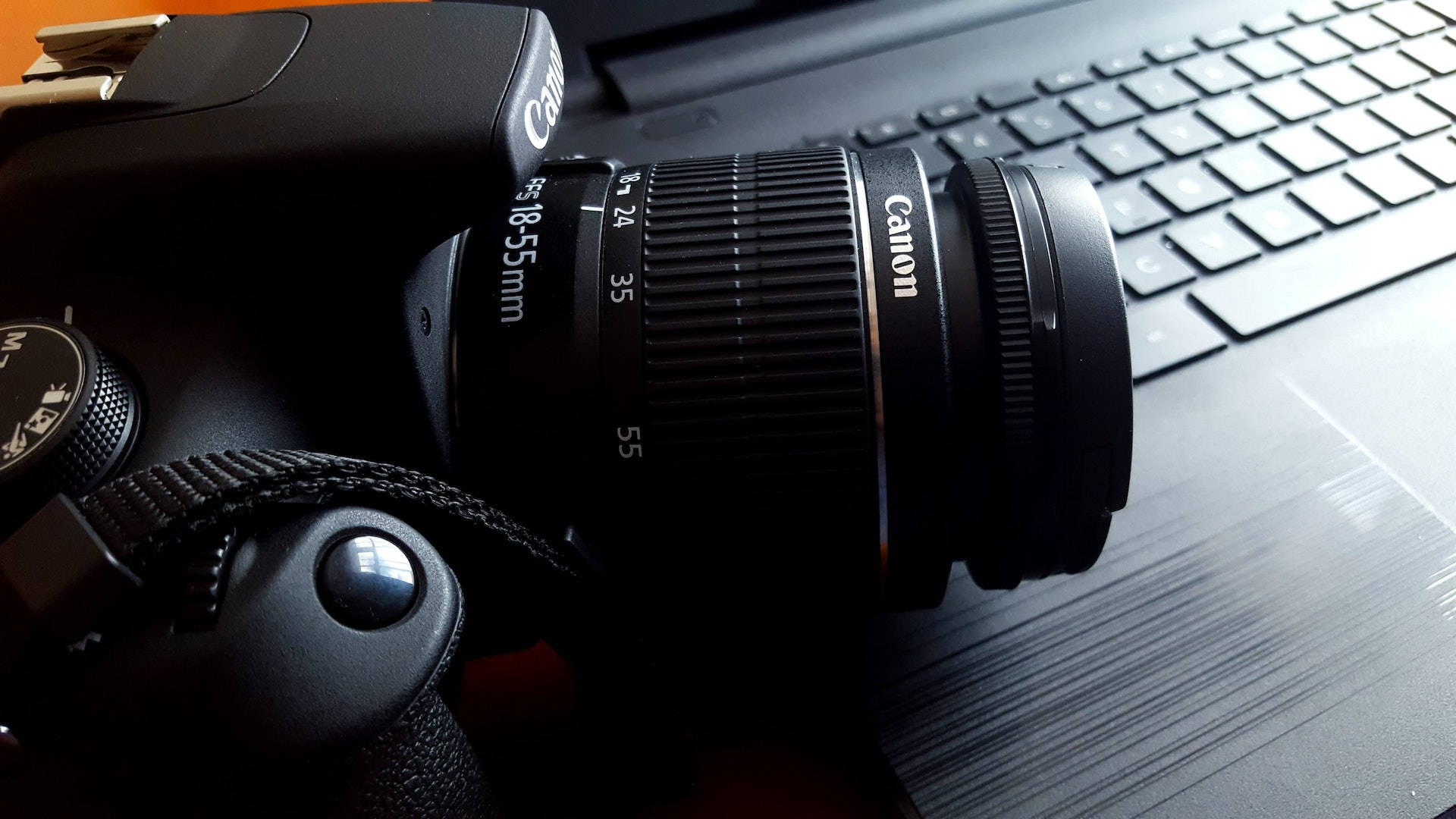 相機 桌布 壁紙 — 一部黑色Canon DSLR相機配備18-55mm鏡頭，放置在昏暗環境中的筆記本電腦鍵盤上。