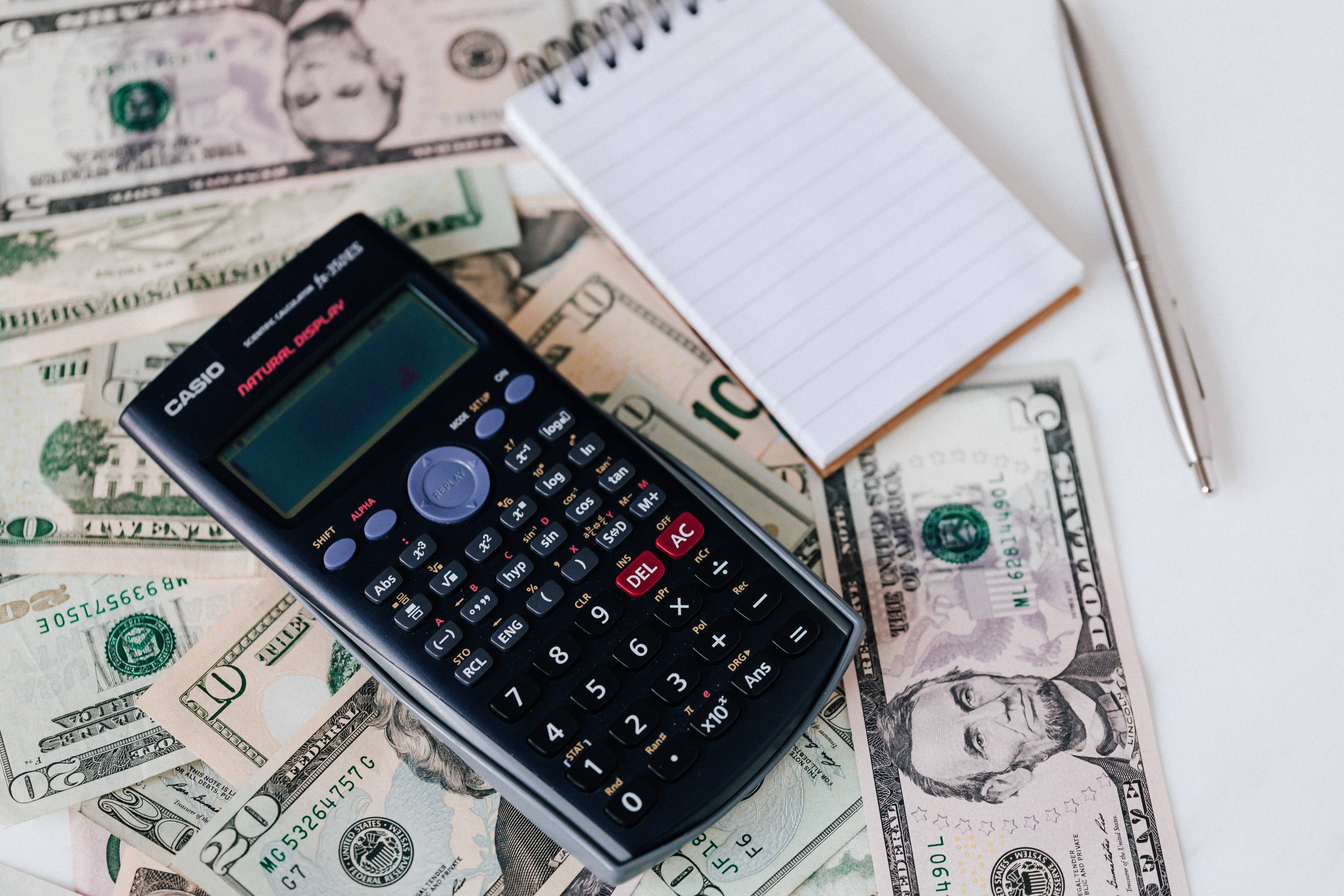 Chartered Accountant 桌布 壁紙 — 一部黑色Casio計算機放在散落的美鈔上，附近有螺旋筆記本和筆。