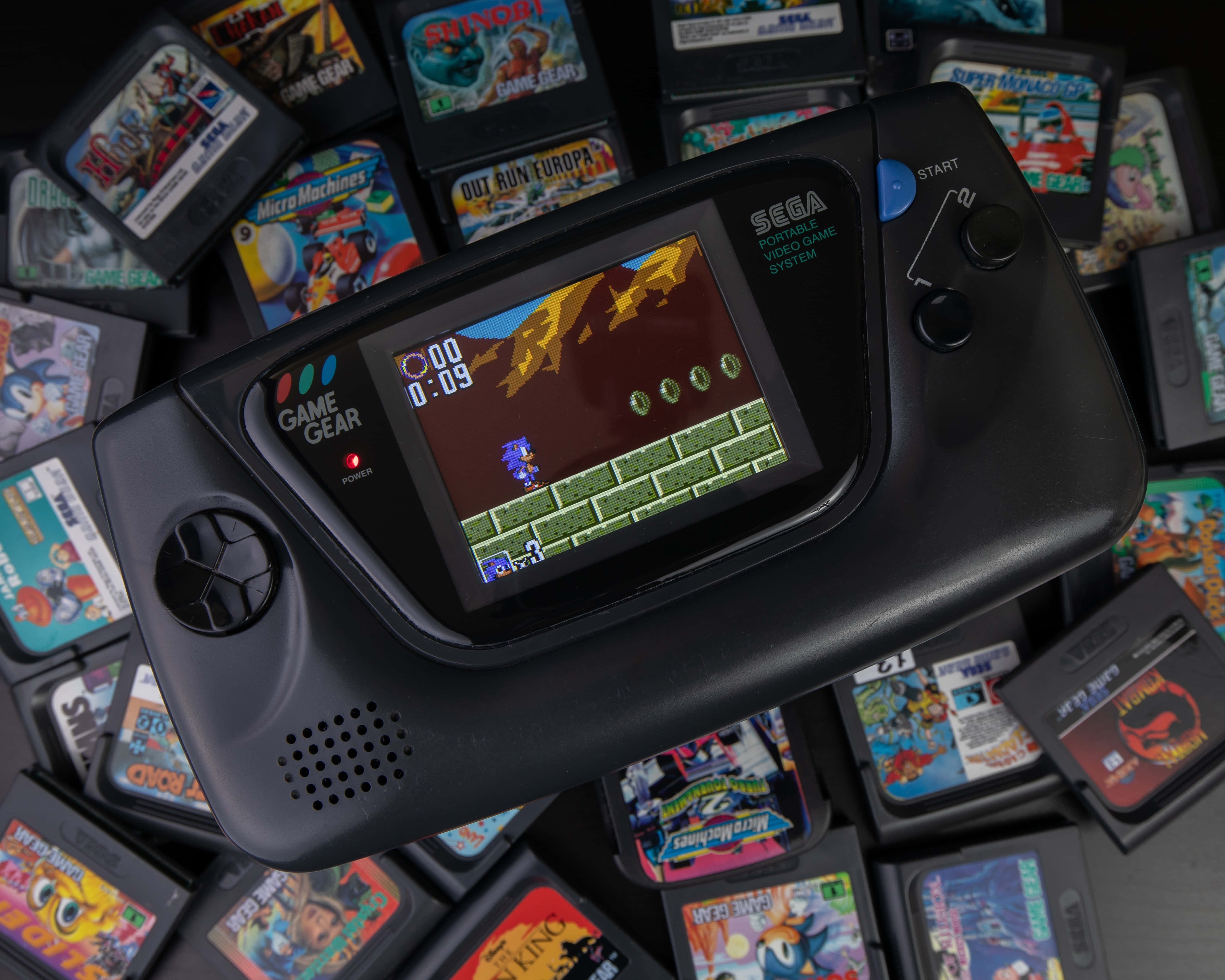Gaming Consoles 桌布 壁紙 — 一部黑色Sega Game Gear手提遊戲機顯示著Sonic遊戲，周圍環繞著色彩繽紛的遊戲卡帶。