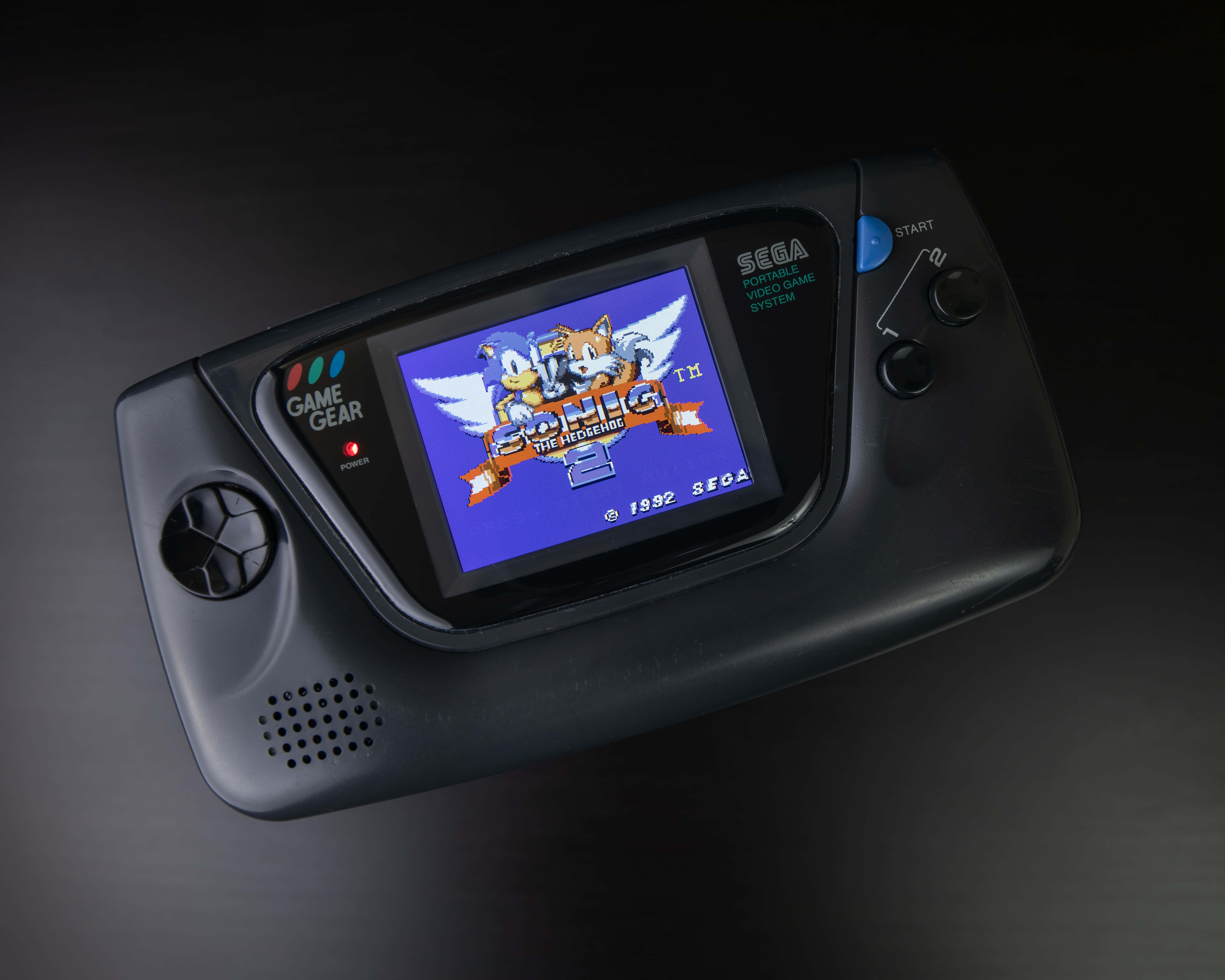 Gaming Consoles 桌布 壁紙 — 一部黑色Sega Game Gear手提遊戲機在深色反光表面上顯示Sonic the Hedgehog 2的標題畫面。