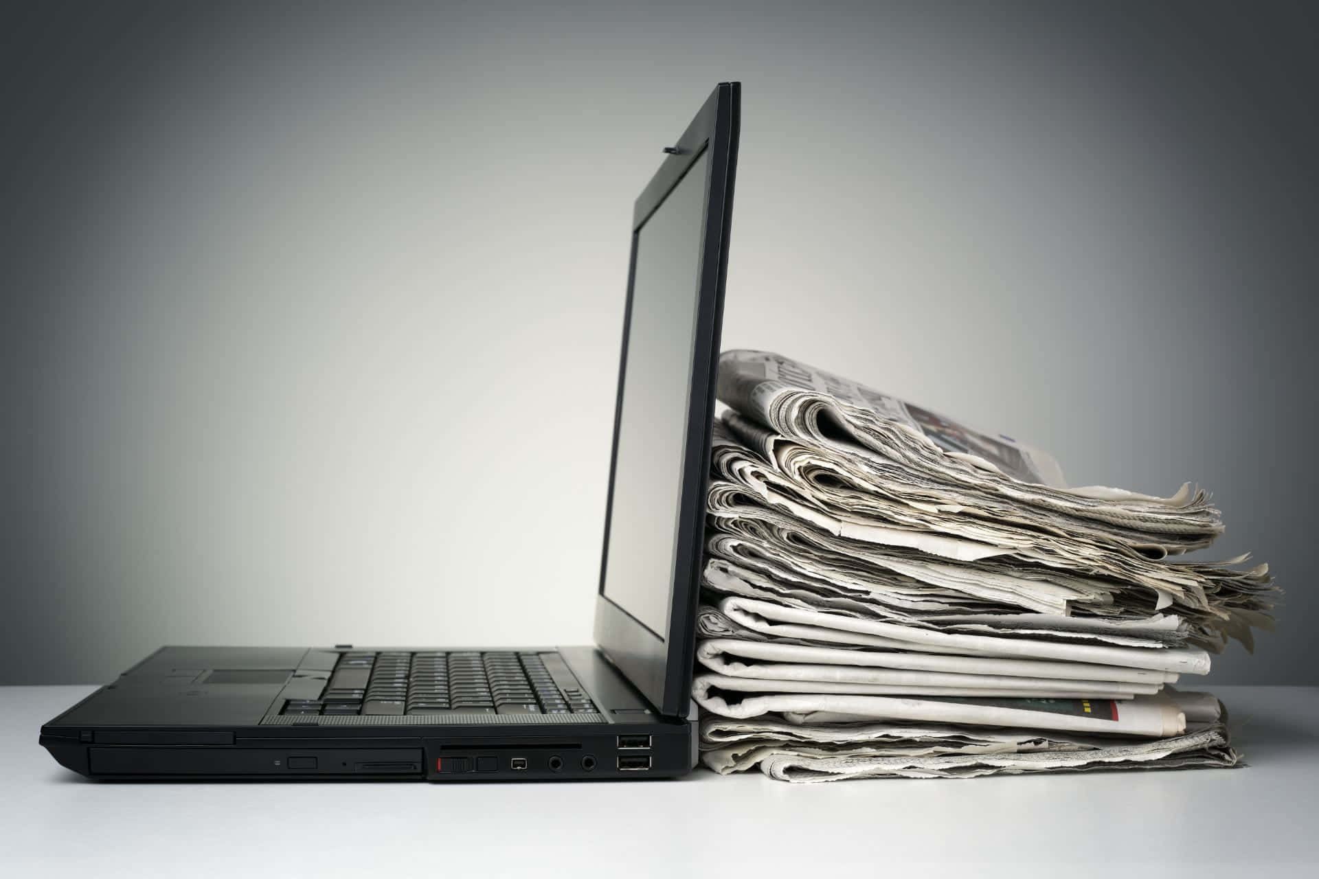 Journalism 桌布 壁紙 — 一部黑色手提電腦直立靠在一疊摺疊報紙上，置於白色表面上。