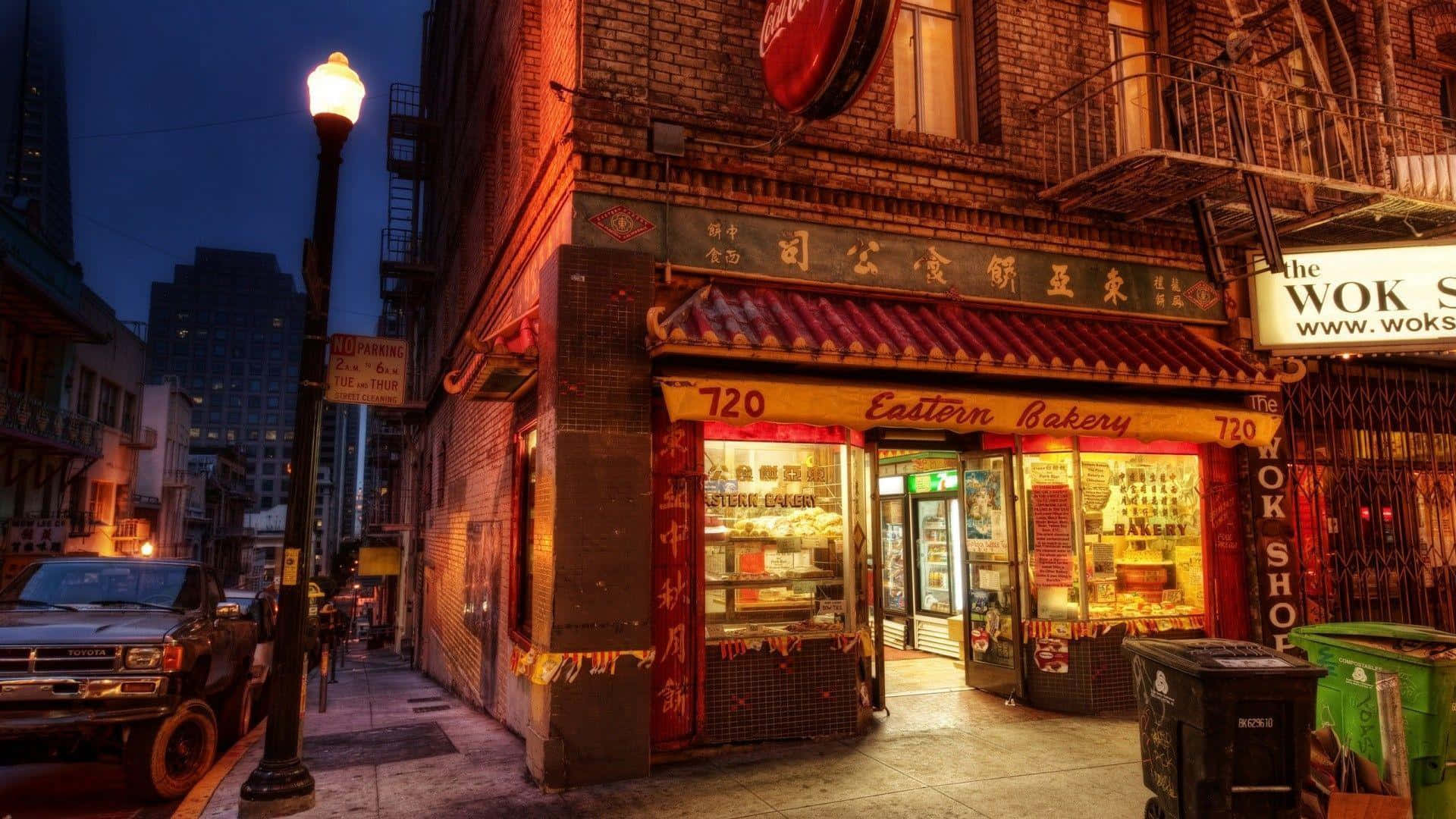 Bakery 桌布 壁紙 — 一間Eastern Bakery的店面在黃昏時分溫暖地發光，位於磚砌的城市小巷中，有一輛停泊的貨車和發光的街燈。