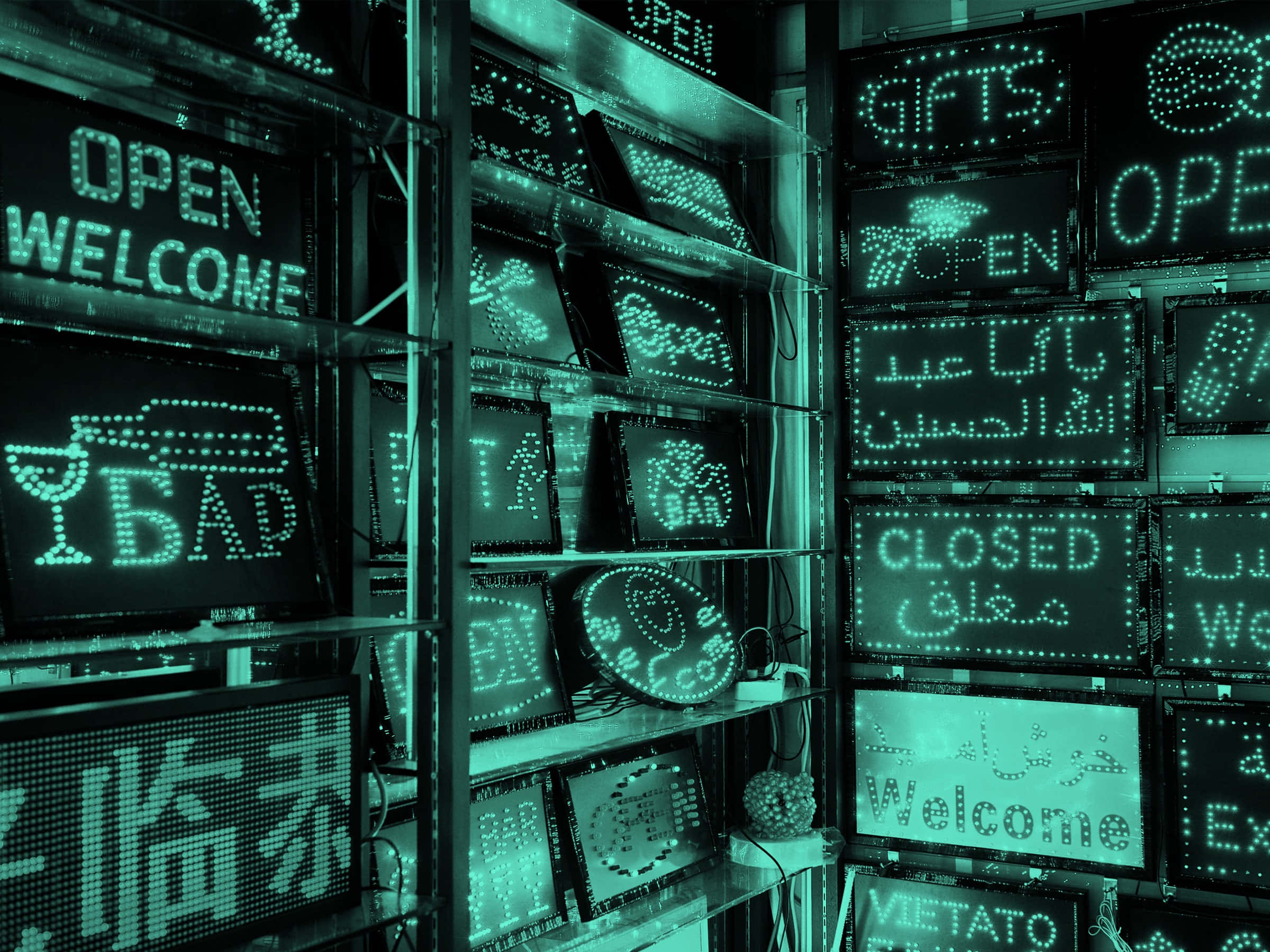 程式碼 桌布 壁紙 — 一間以青綠色燈光照亮的商店展示著多層架的多語言LED標誌，上面有各種文字的「OPEN」「WELCOME」和「CLOSED」訊息。