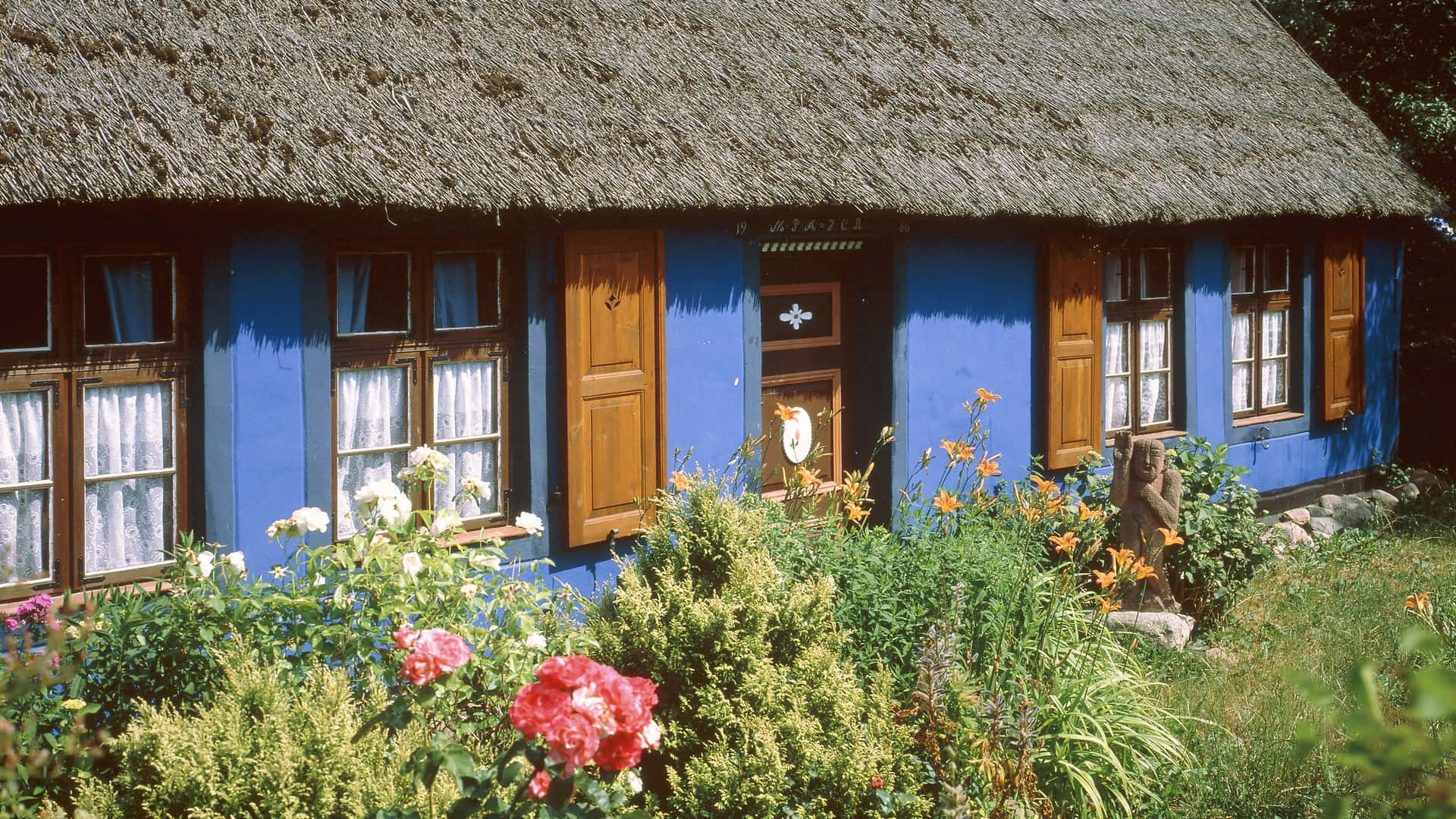 Cottage Garden 桌布 壁紙 — 一間充滿活力的藍色茅草屋頂小屋，配有木製百葉窗，坐落在一個茂盛的花園中，在明亮的陽光下綻放著色彩繽紛的花朵。