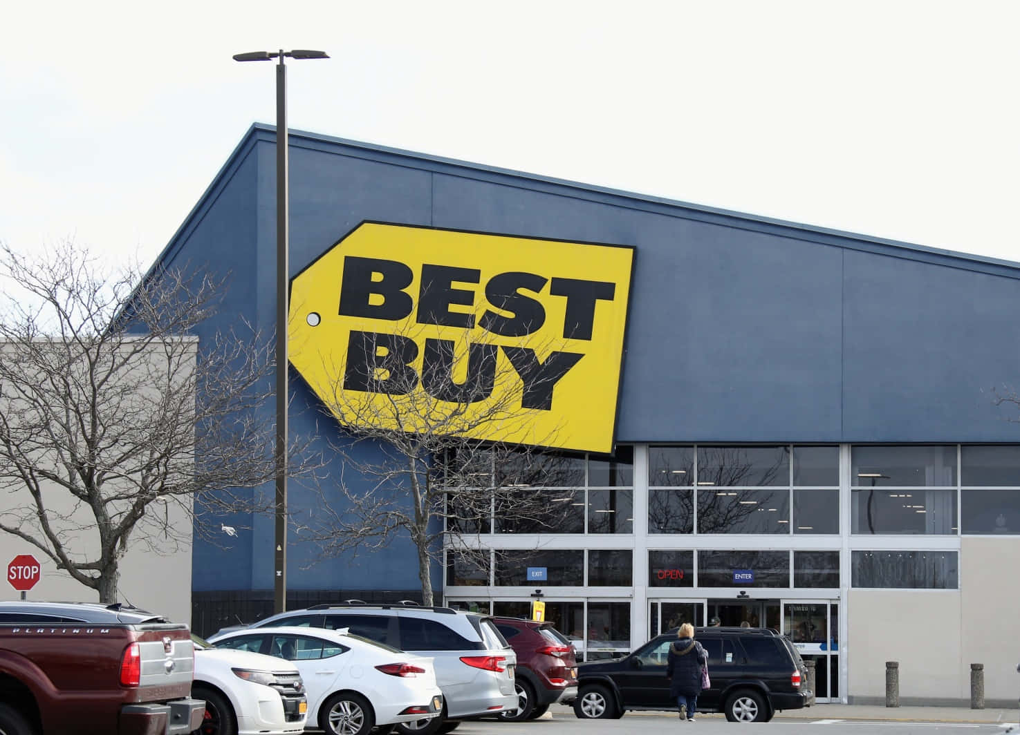 buy 桌布 壁紙 — 一間大型藍色Best Buy商店，帶有亮黃色標誌，矗立在陰天之下，汽車正在停車，一個人正走向入口。