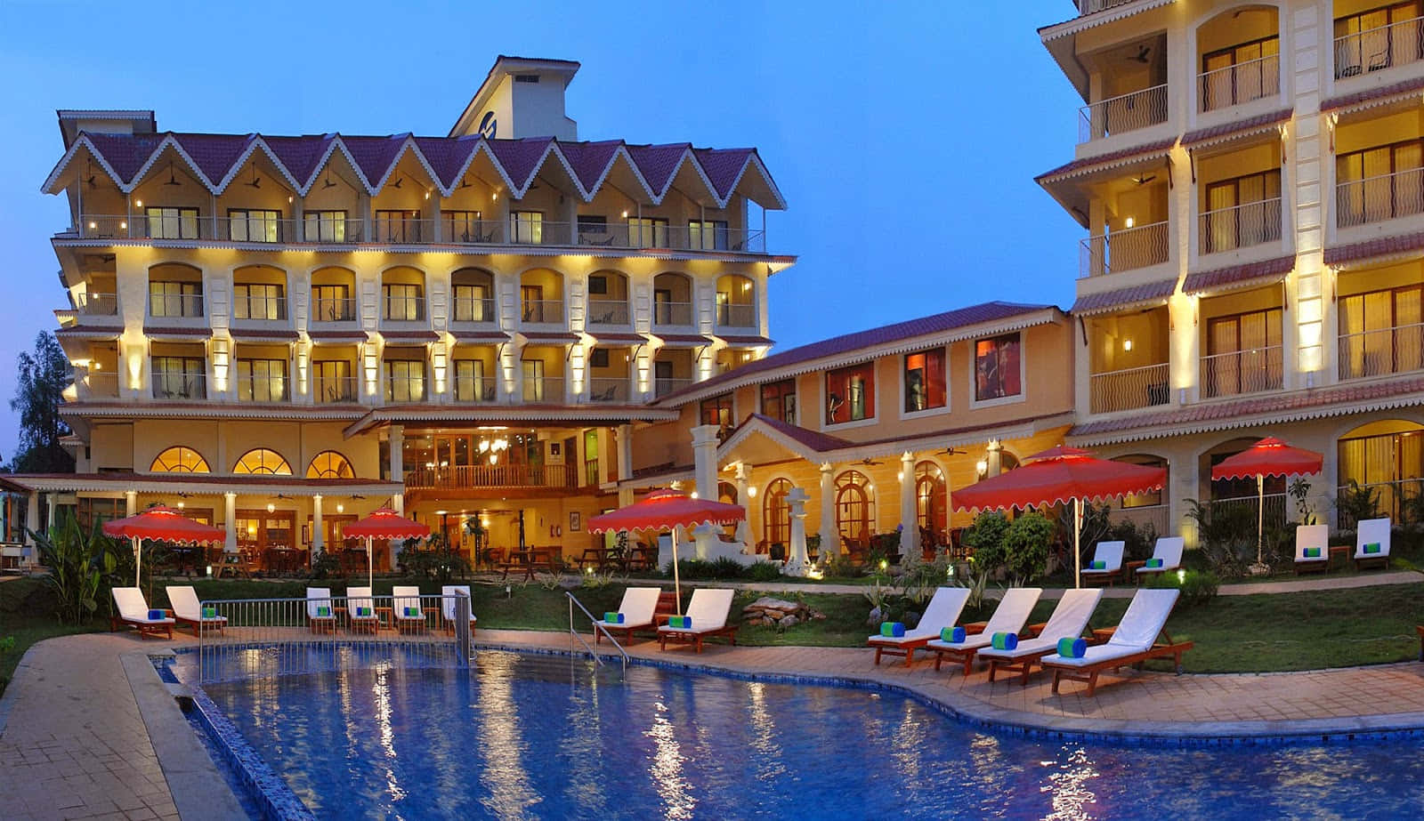 Hotel 桌布 壁紙 — 一間宏偉的酒店，溫暖的燈光和紅色遮陽傘環繞著暮色中寧靜的泳池。