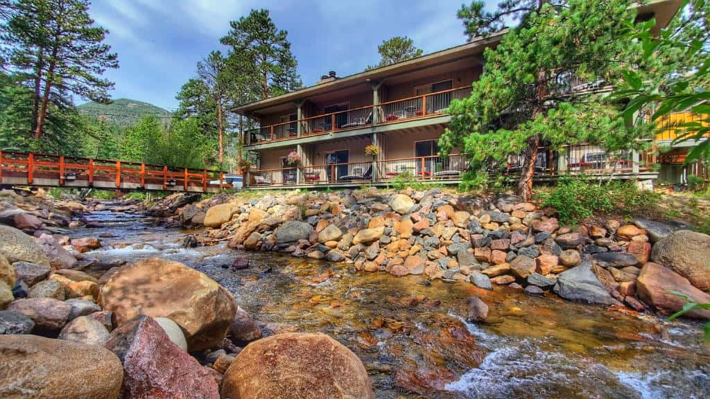 Fall Cabin 桌布 壁紙 — 一間帶陽台的木屋坐落在岩石溪流旁，溪流在紅橋下流過，四周環繞著高大的松樹。