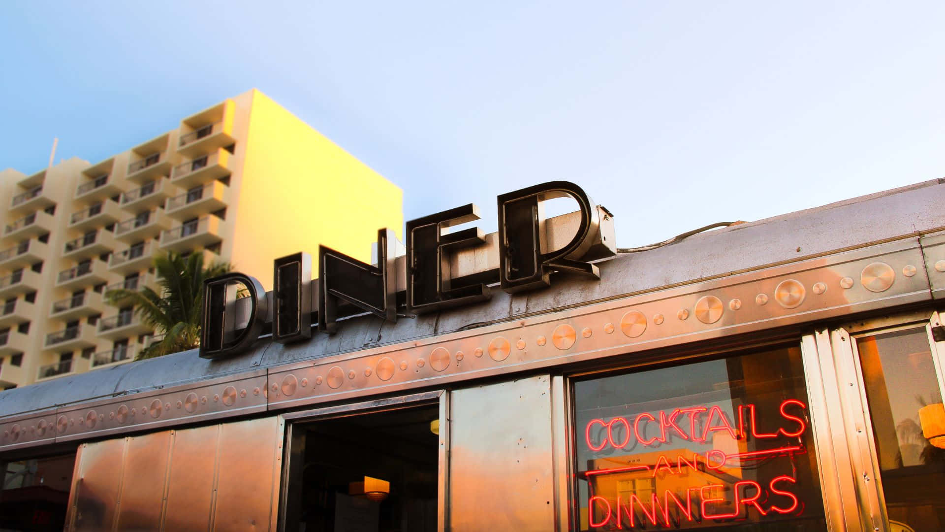 Diner 桌布 壁紙 — 一間復古風格的餐廳，帶有醒目的黑色招牌和發光的紅色霓虹燈雞尾酒和晚餐招牌，襯托著溫暖的日落天空。