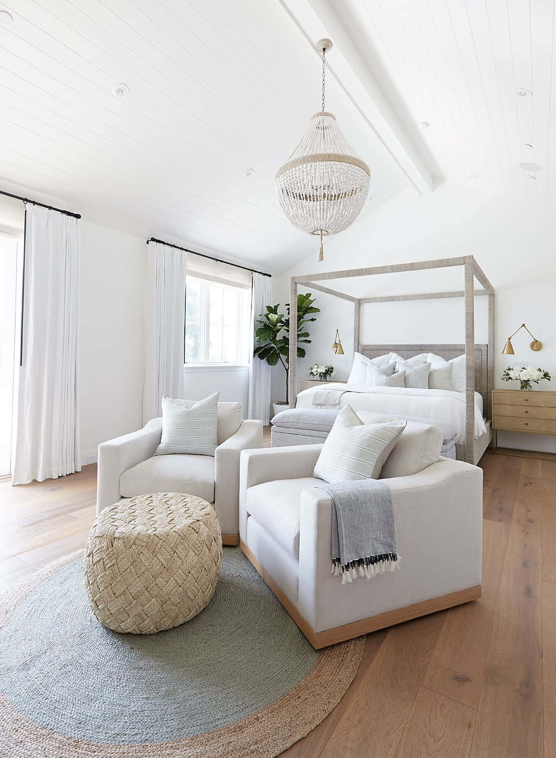 Interior Design 桌布 壁紙 — 一間明亮通風的臥室，配有白色護牆板、木地板和一張四柱床，周圍環繞著舒適的白色扶手椅和編織腳凳，上方懸掛著水晶吊燈。