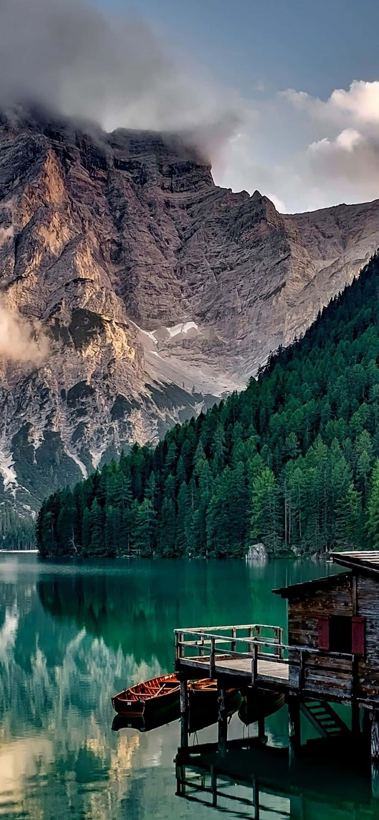 Mountain iPhone 桌布 壁紙 — 一間木屋坐落在碼頭上，旁邊是清澈碧綠的湖泊，倒映著高聳霧濛濛的山脈和茂密的松樹林。