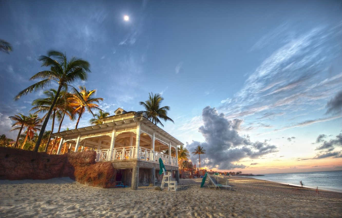 Bahamas Beach 桌布 壁紙 — 一間海濱小屋，配有白色欄杆和棕櫚樹，在暮色天空下閃耀，天空中有明亮的月亮和柔軟的雲朵。