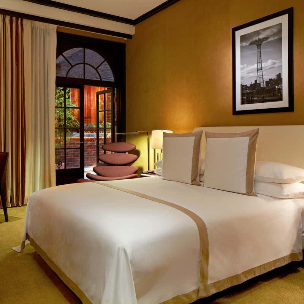 Hotel Room 桌布 壁紙 — 一間溫暖照明的酒店睡房，配有整齊鋪好的床、通往庭院的拱形玻璃門，以及芥末黃牆上裱框的城市景觀照片。