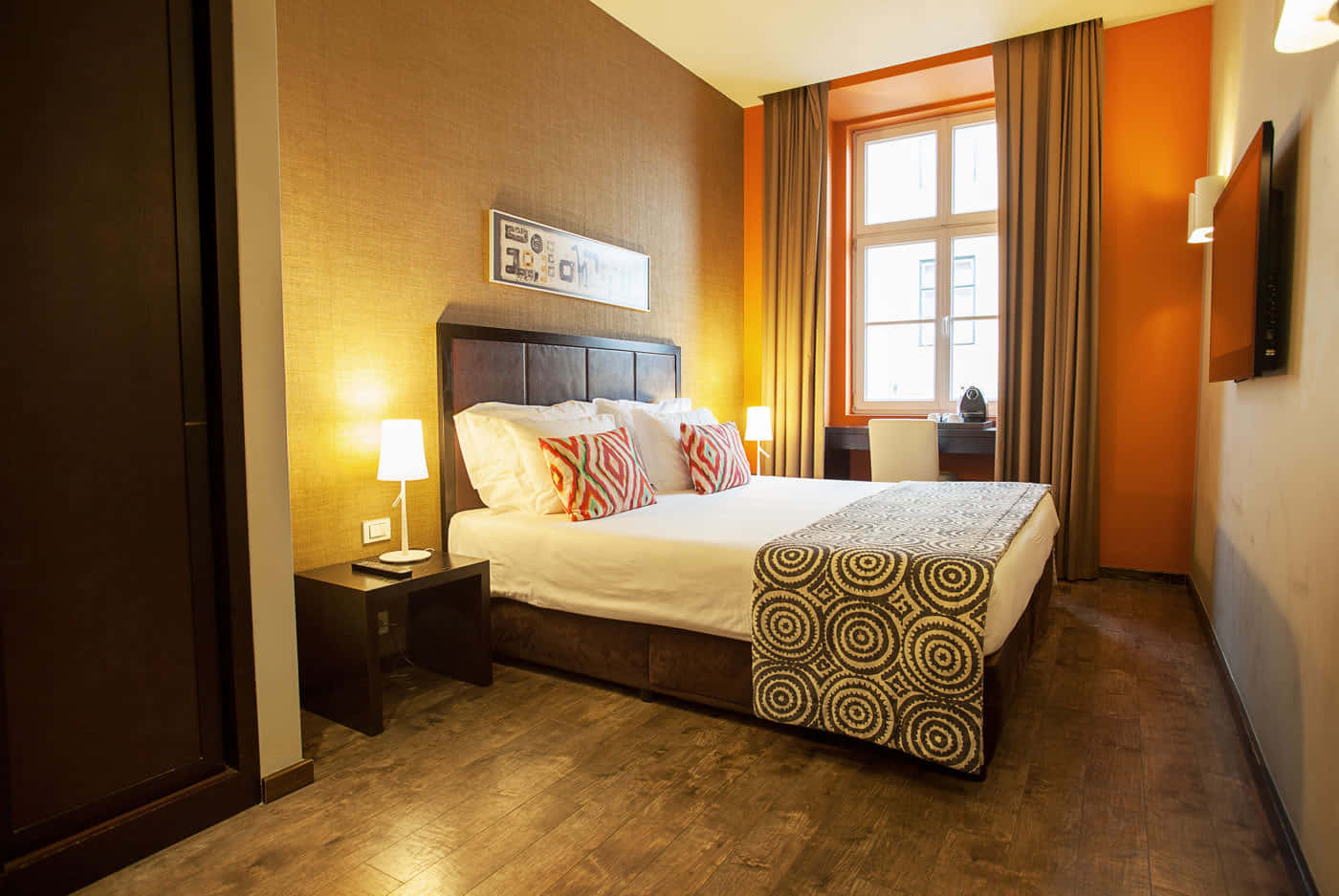 Hotel Room 桌布 壁紙 — 一間燈光溫暖的酒店睡房，設有橙色牆壁、木地板，以及一張飾有圖案枕頭和螺旋毯子的床。