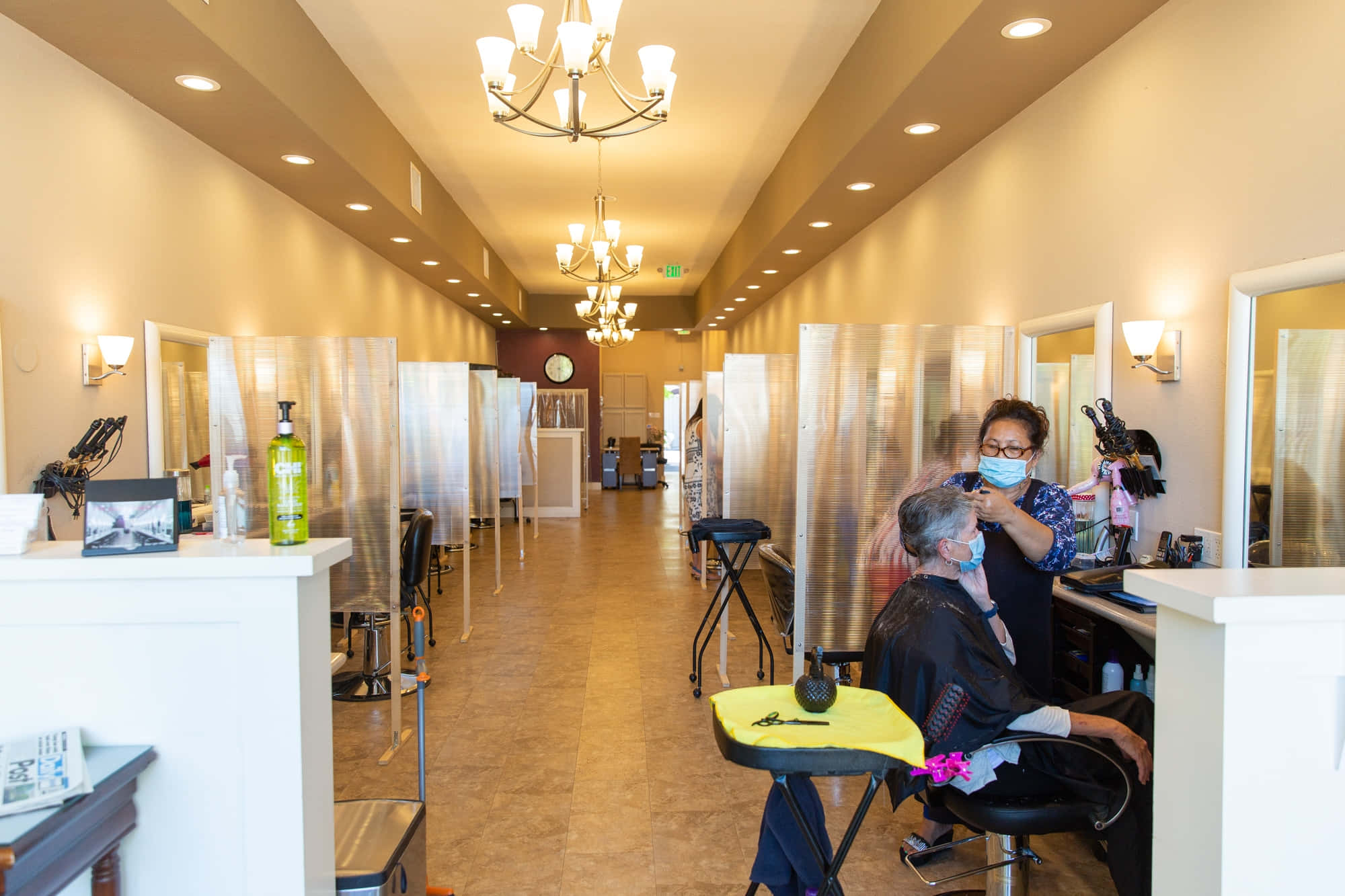 Hair Salon 桌布 壁紙 — 一間燈光溫暖的髮廊，造型師戴着口罩，在透明膠隔板後面為客人剪髮。