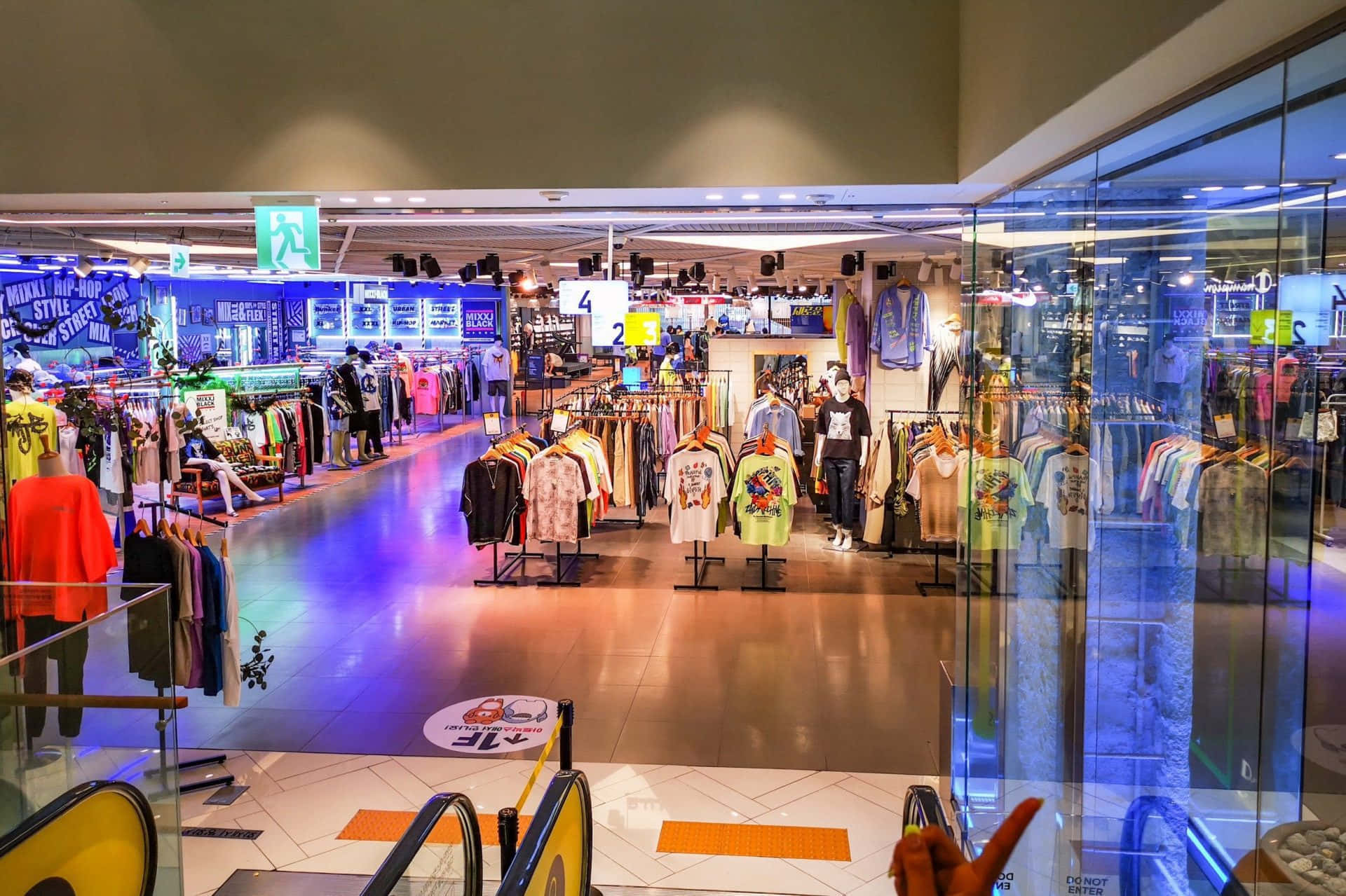 Mall 桌布 壁紙 — 一間燈火通明的服裝店，高天花板下陳列著色彩繽紛的襯衫架和發光的藍色霓虹招牌。