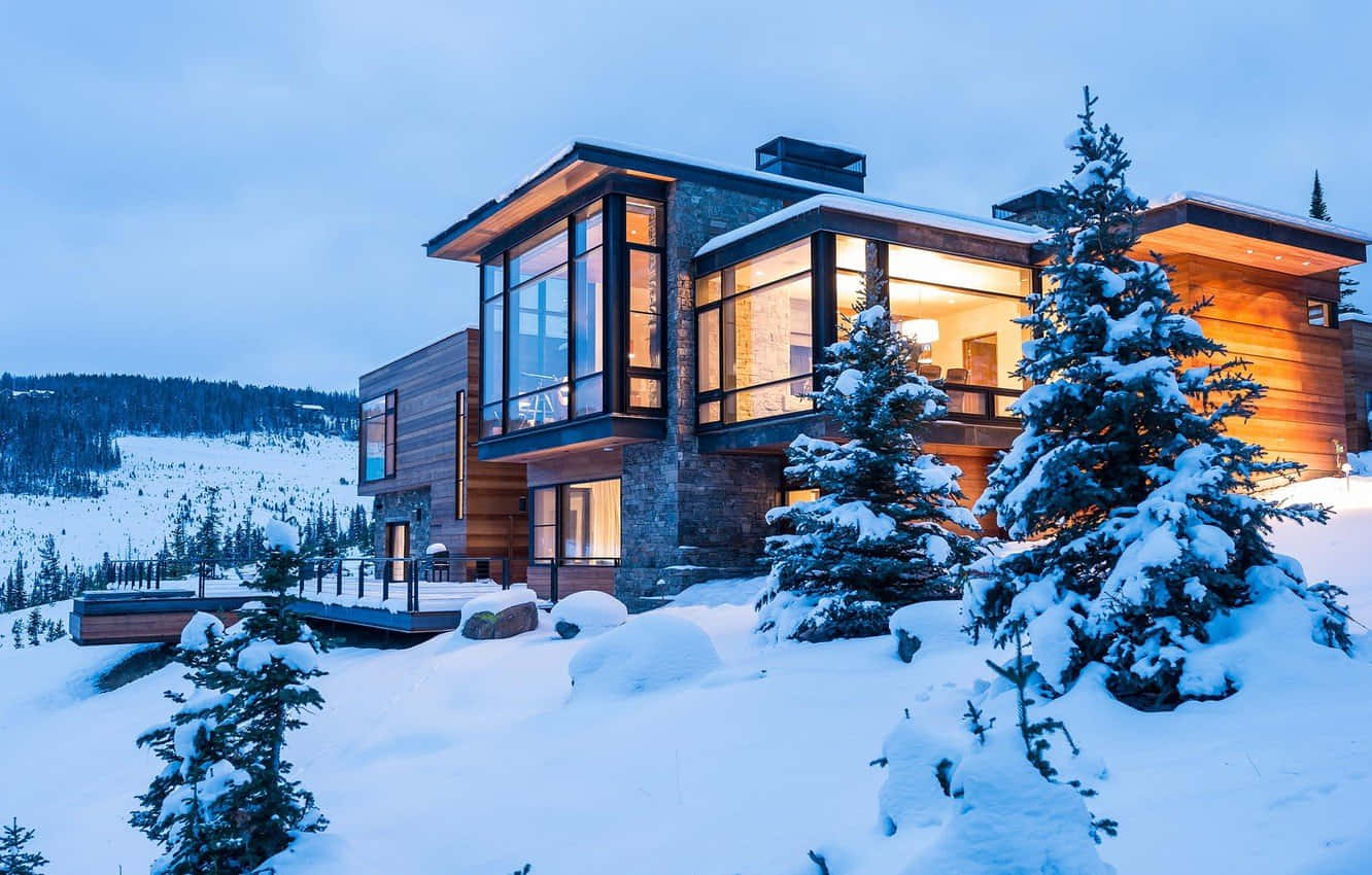 Cozy Winter Cabin 桌布 壁紙 — 一間現代木屋，窗戶閃爍著光芒，坐落在暮色天空下白雪皚皚的山景中。