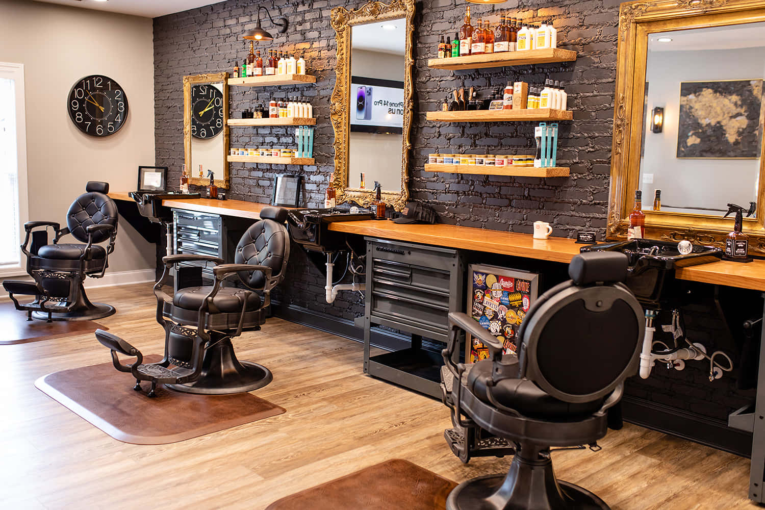 Hair Salon 桌布 壁紙 — 一間現代理髮店，配有黑色皮革椅、木製櫃檯和磚牆，裝飾著金色鏡子和擺放護理產品的架子。