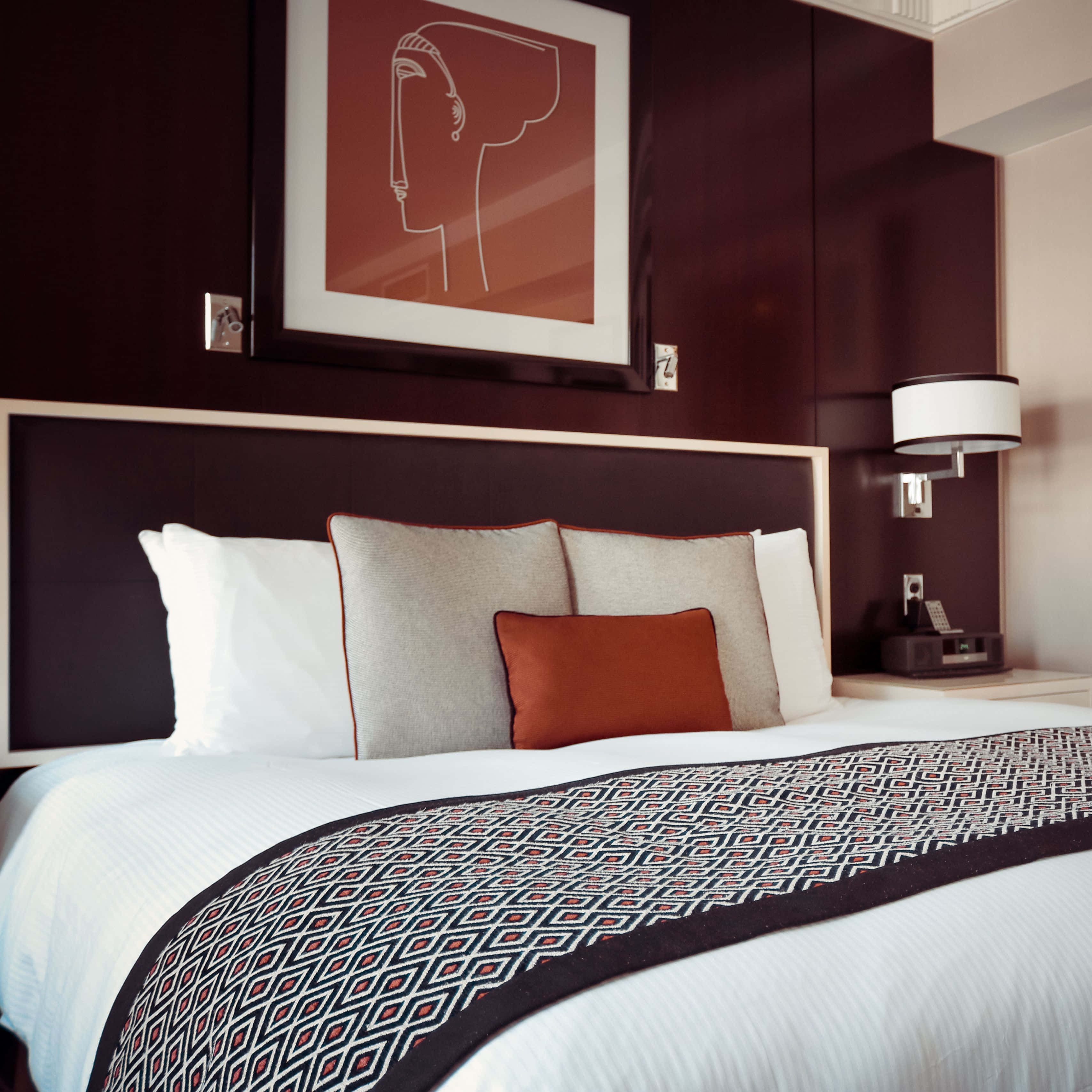 Hotel Room 桌布 壁紙 — 一間現代酒店睡房，床鋪整齊，配有幾何圖案床尾巾，深色床頭板上方掛有抽象藝術品。