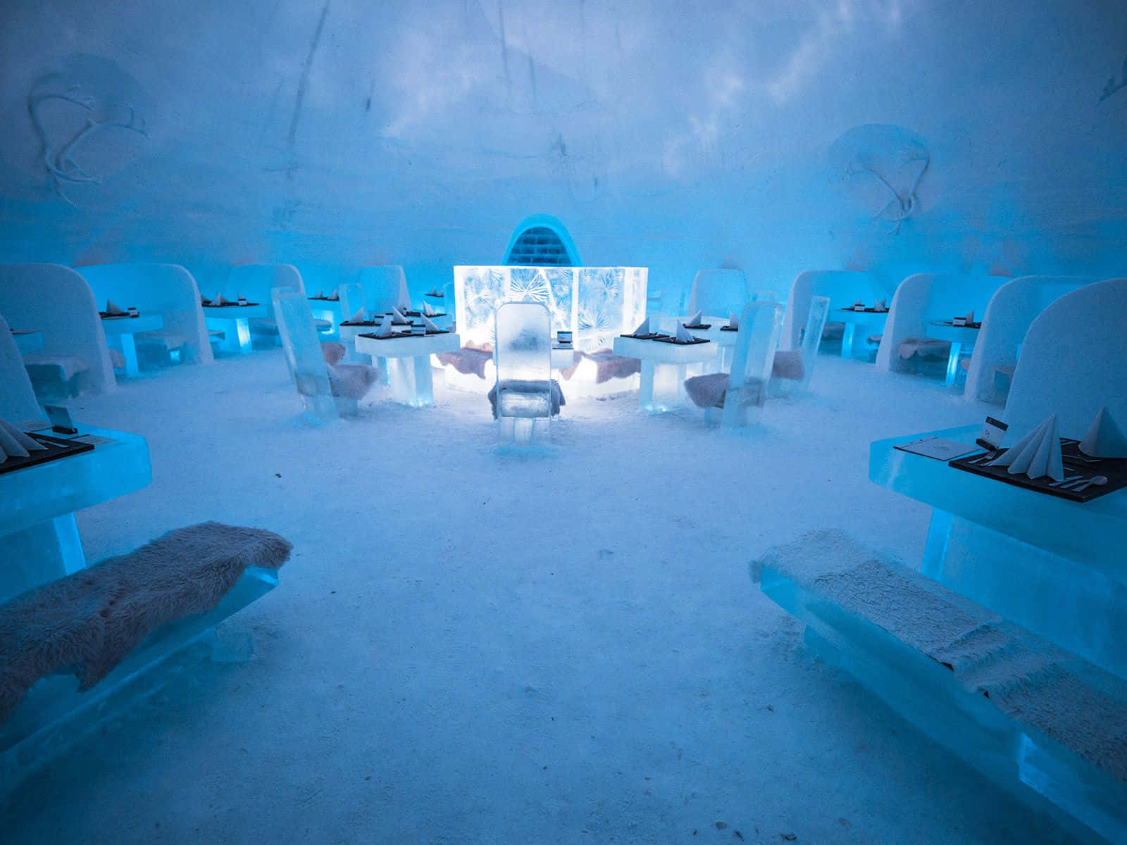 Ice Hotel 桌布 壁紙 — 一間發光的藍色冰餐廳，在彎曲的冰天花下設有雕刻的桌子、椅子和長凳。