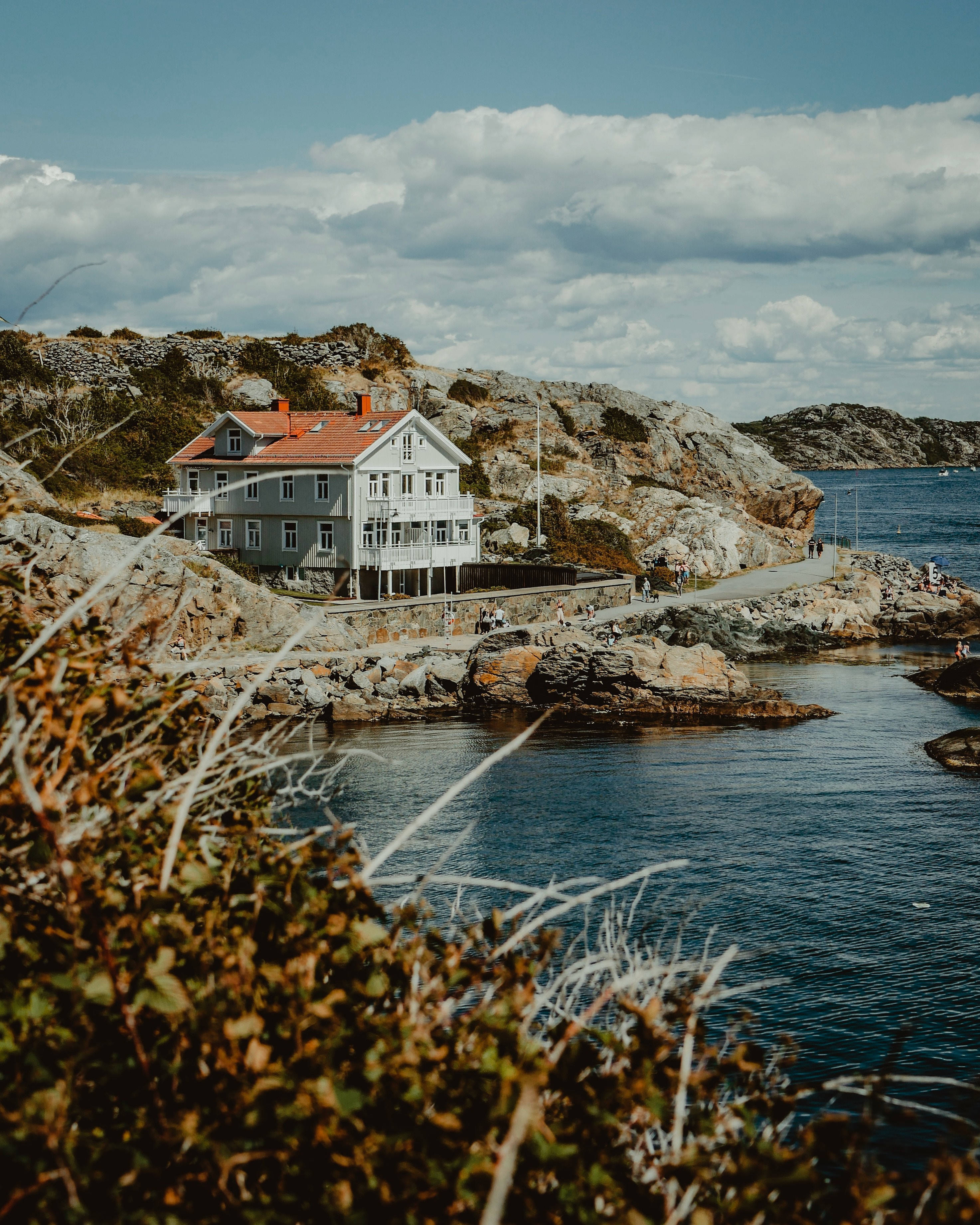 Sweden 桌布 壁紙 — 一間白色海岸房屋，紅色屋頂，棲息在岩石峭壁上，俯瞰深藍色海水，天空部分多雲。