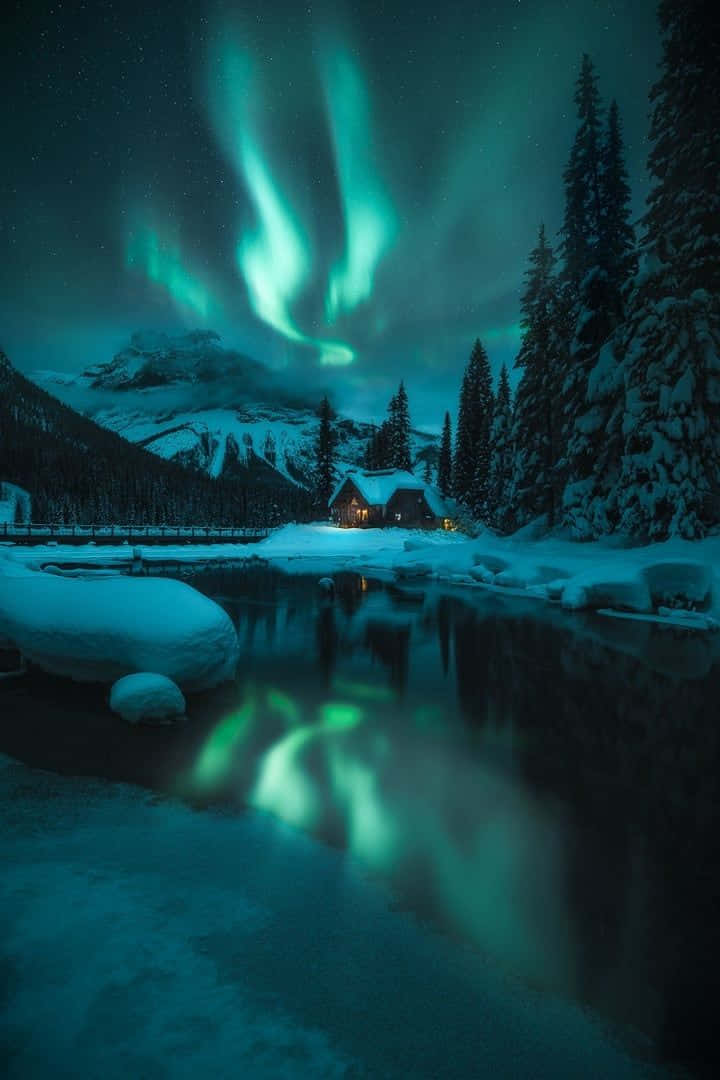 Winter Photography 桌布 壁紙 — 一間舒適的小屋坐落在白雪皚皚的冬季森林中，上方是 vibrant 的綠色北極光，倒映在平靜的河流中。