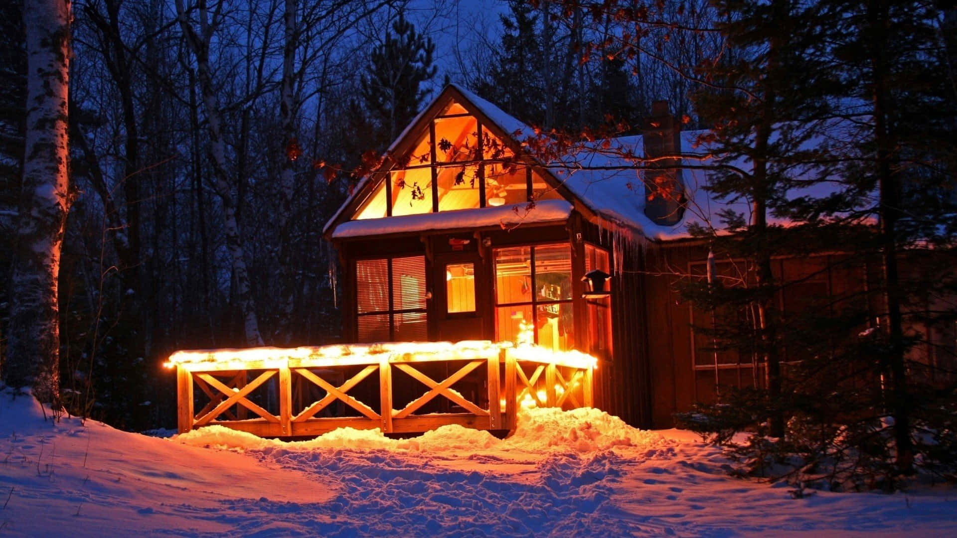 Cozy Winter Cabin 桌布 壁紙 — 一間舒適的小屋坐落在白雪皚皚的樹林中，內部散發著溫暖的光芒，而暮色將天空染成深藍色。