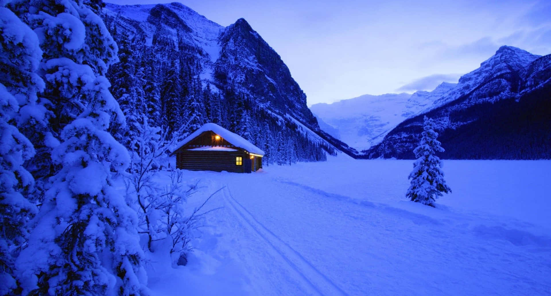 Cozy Winter Cabin 桌布 壁紙 — 一間舒適的木屋在暮色中，對著廣闊的雪山山谷溫暖地發光。