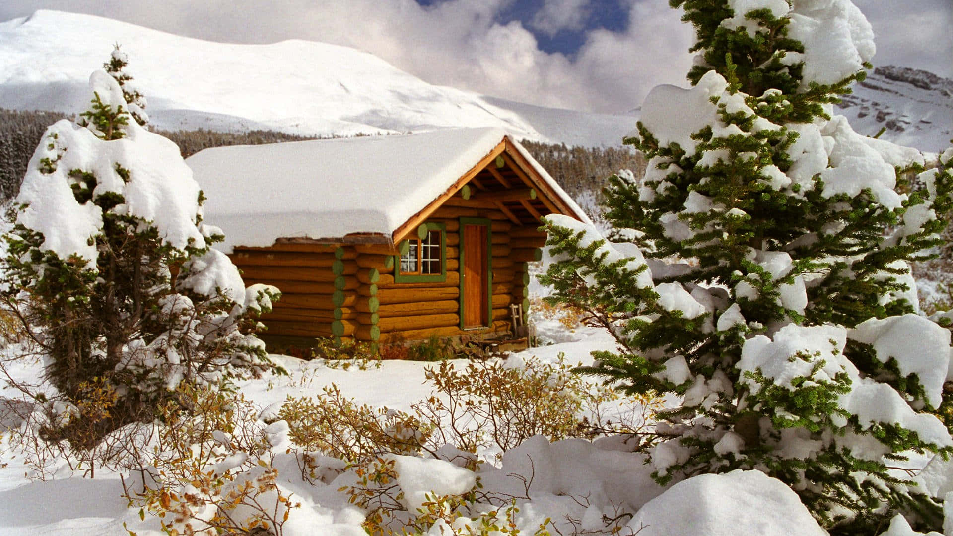Cozy Winter Cabin 桌布 壁紙 — 一間舒適的木屋坐落在白雪皚皚的山景中，常青樹上積滿了雪，天空湛藍明亮。