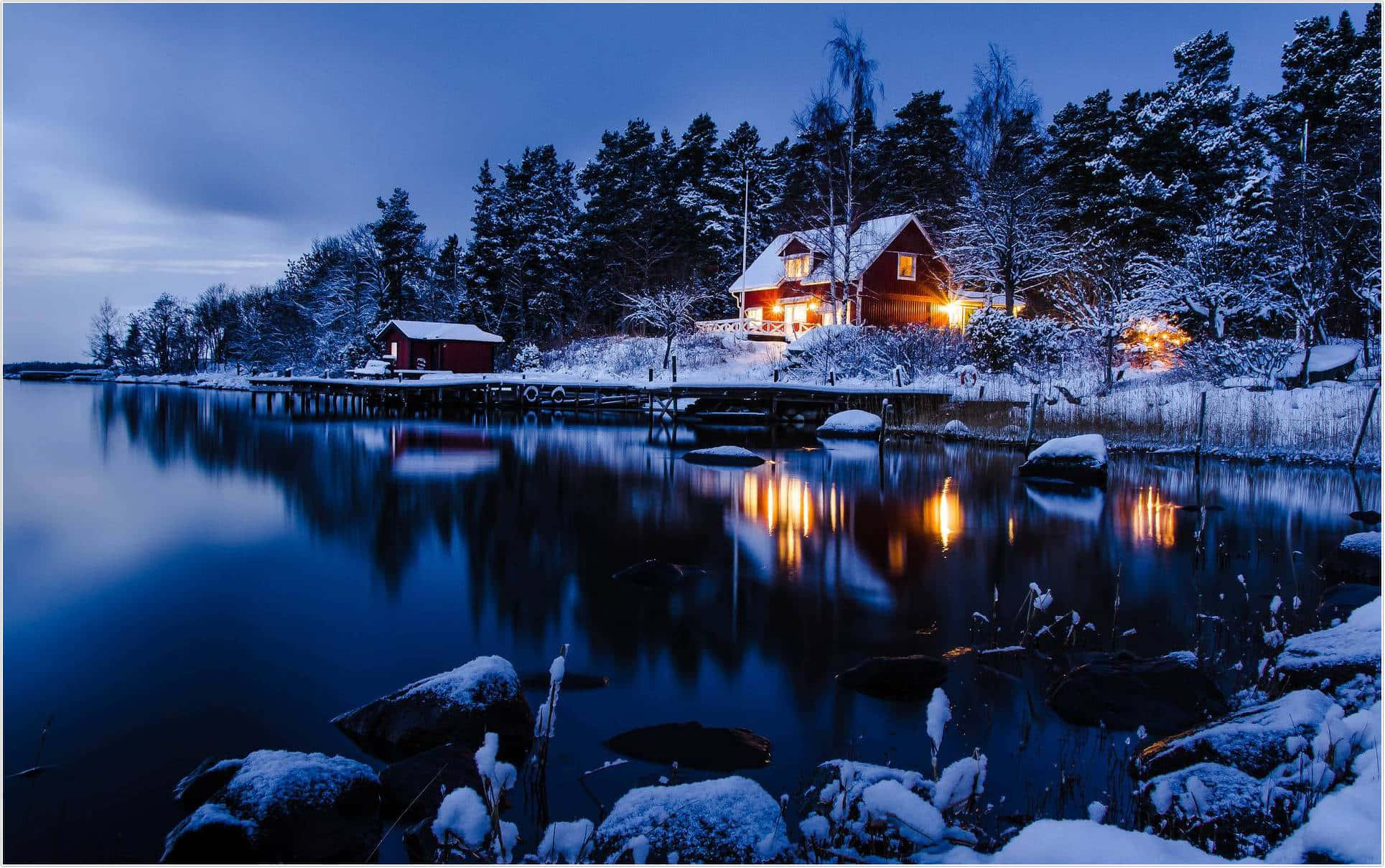 Winter Blues 桌布 壁紙 — 一間舒適的紅色小屋，溫暖的燈光在暮色中映照著白雪覆蓋的森林，平靜的冰湖上閃爍著它的倒影。
