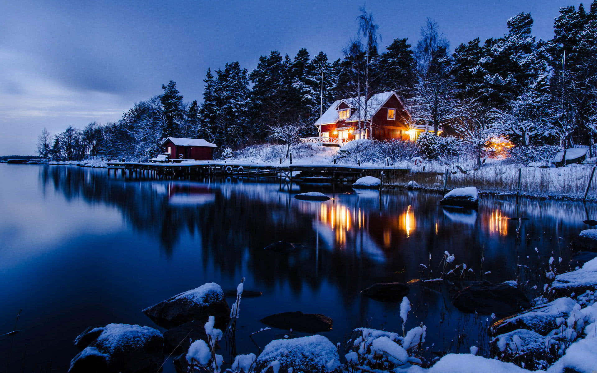 冬季場景 桌布 壁紙 — 一間舒適的紅色小屋在雪景暮色湖邊溫暖地發光，燈光倒映在平靜的水面上，周圍是白雪覆蓋的樹木。
