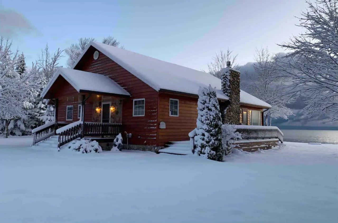 Cozy Winter Cabin 桌布 壁紙 — 一間舒適的紅色木屋被雪覆蓋，坐落在結冰的湖邊，黃昏時窗戶透出溫暖的燈光。