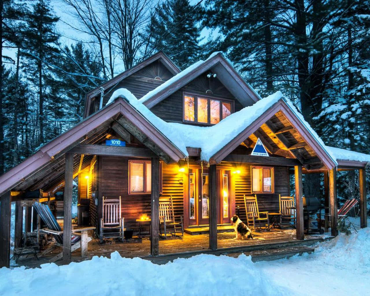 Cozy Winter Cabin 桌布 壁紙 — 一間舒適的雪覆蓋小屋在黃昏時亮著燈，溫暖地發光，周圍是高聳的松樹，一隻狗坐在門廊上。