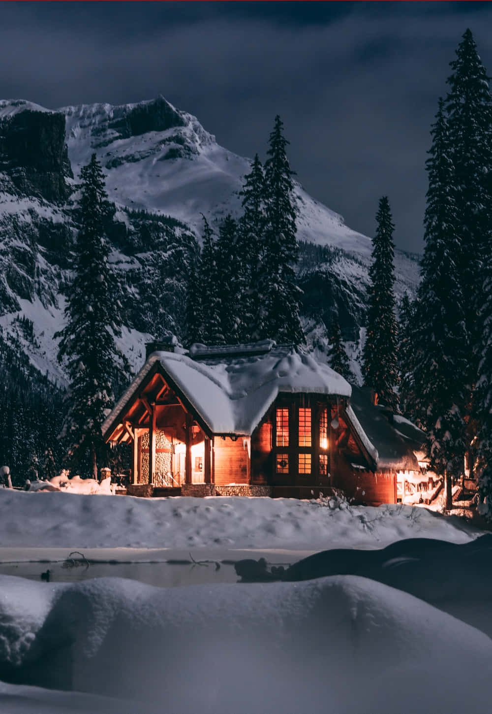 Cozy Winter Cabin 桌布 壁紙 — 一間舒適的雪覆蓋小屋在黑暗的山景背景下，在雪夜天空下溫暖地發光。