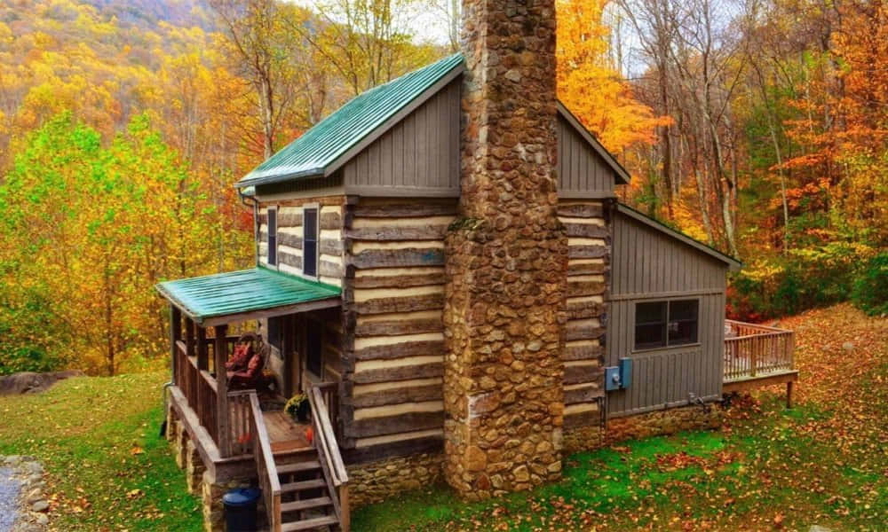Fall Cabin 桌布 壁紙 — 一間質樸的木屋，配有石頭煙囪，坐落在充滿活力的秋日森林中，金色樹葉環繞。