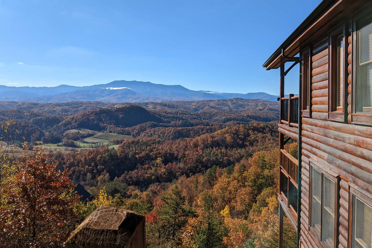Fall Cabin 桌布 壁紙 — 一間質樸的木屋俯瞰著廣闊的秋日山谷，山谷中有起伏的丘陵和遠處白雪覆蓋的山峰，天空湛藍清澈。