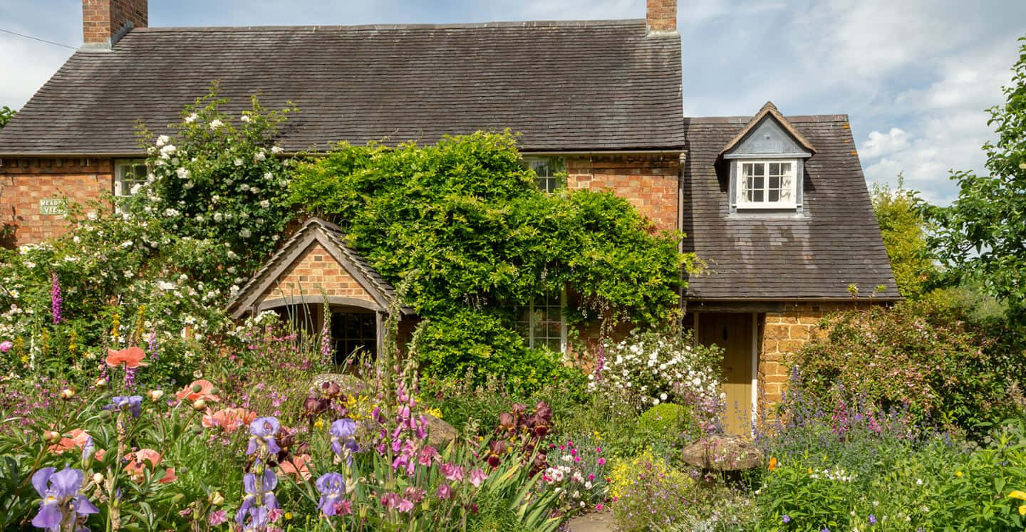 Cottage Garden 桌布 壁紙 — 一間迷人的磚砌小屋，坐落在充滿活力的花園中，花園裡開滿了色彩繽紛的花朵，綠意盎然，天空明亮。