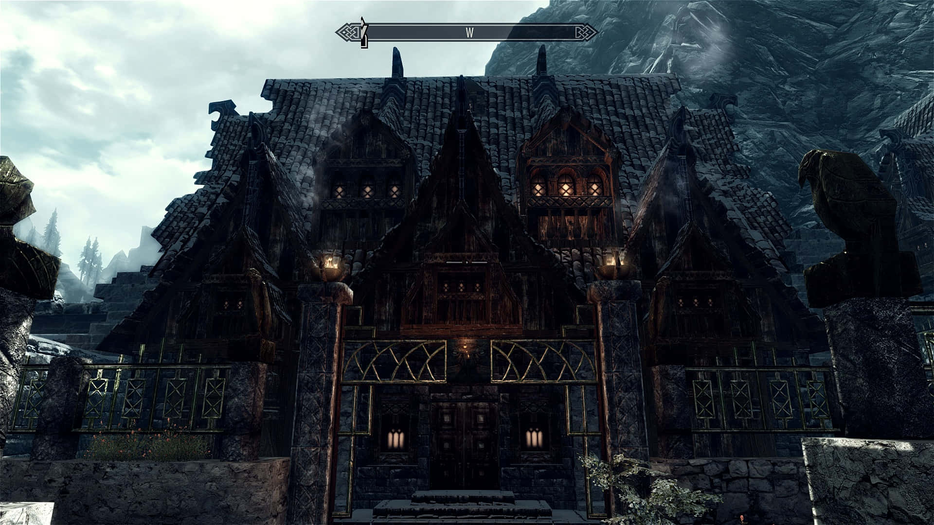 Skyrim Windhelm 桌布 壁紙 — 一間黑暗的中世紀風格房屋，帶有發光的窗戶和石像，矗立在多雲天空下的霧氣山景中。